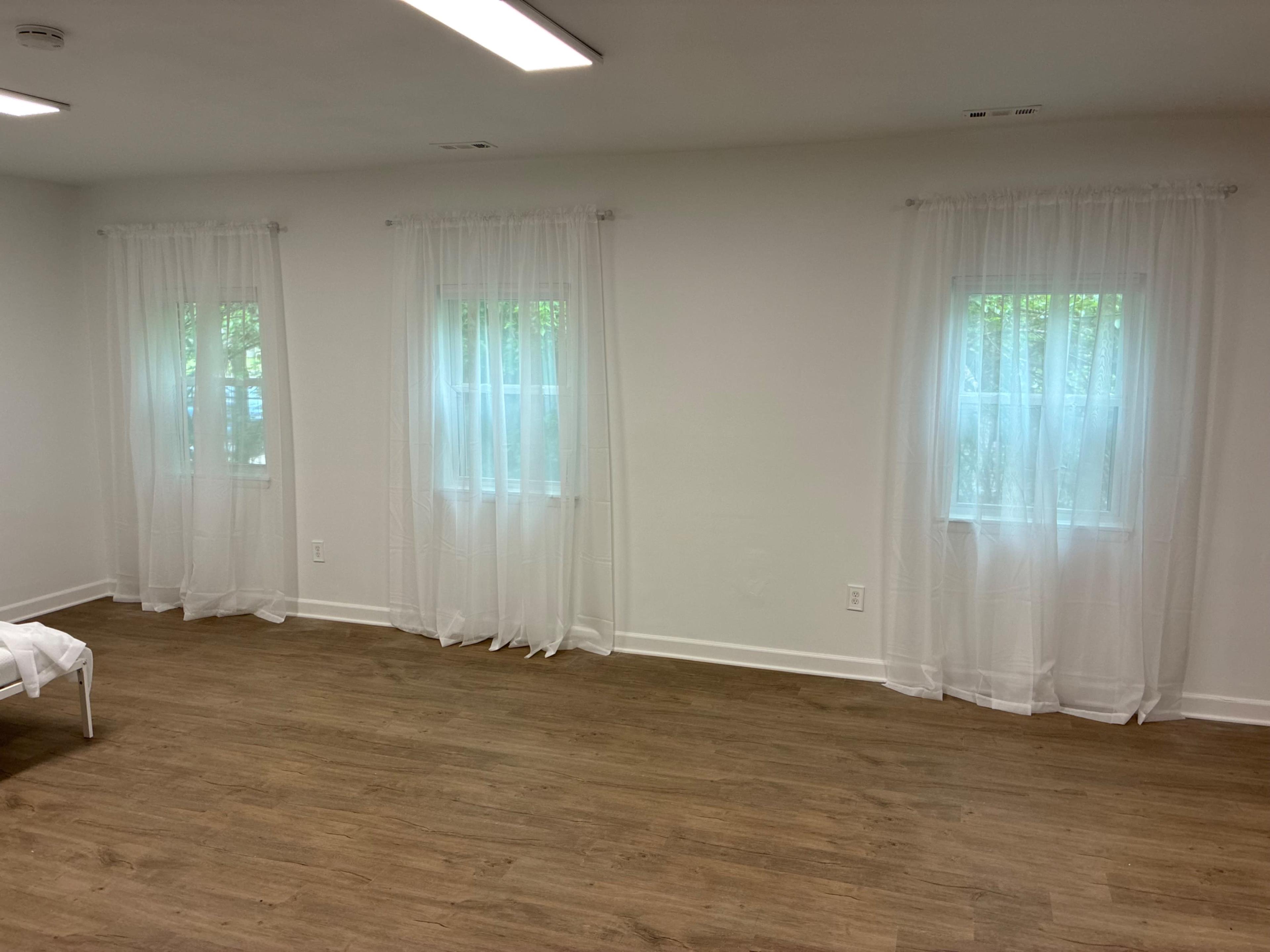 The image shows a room with two windows dressed in sheer white curtains, a light wooden floor, and plain white walls.