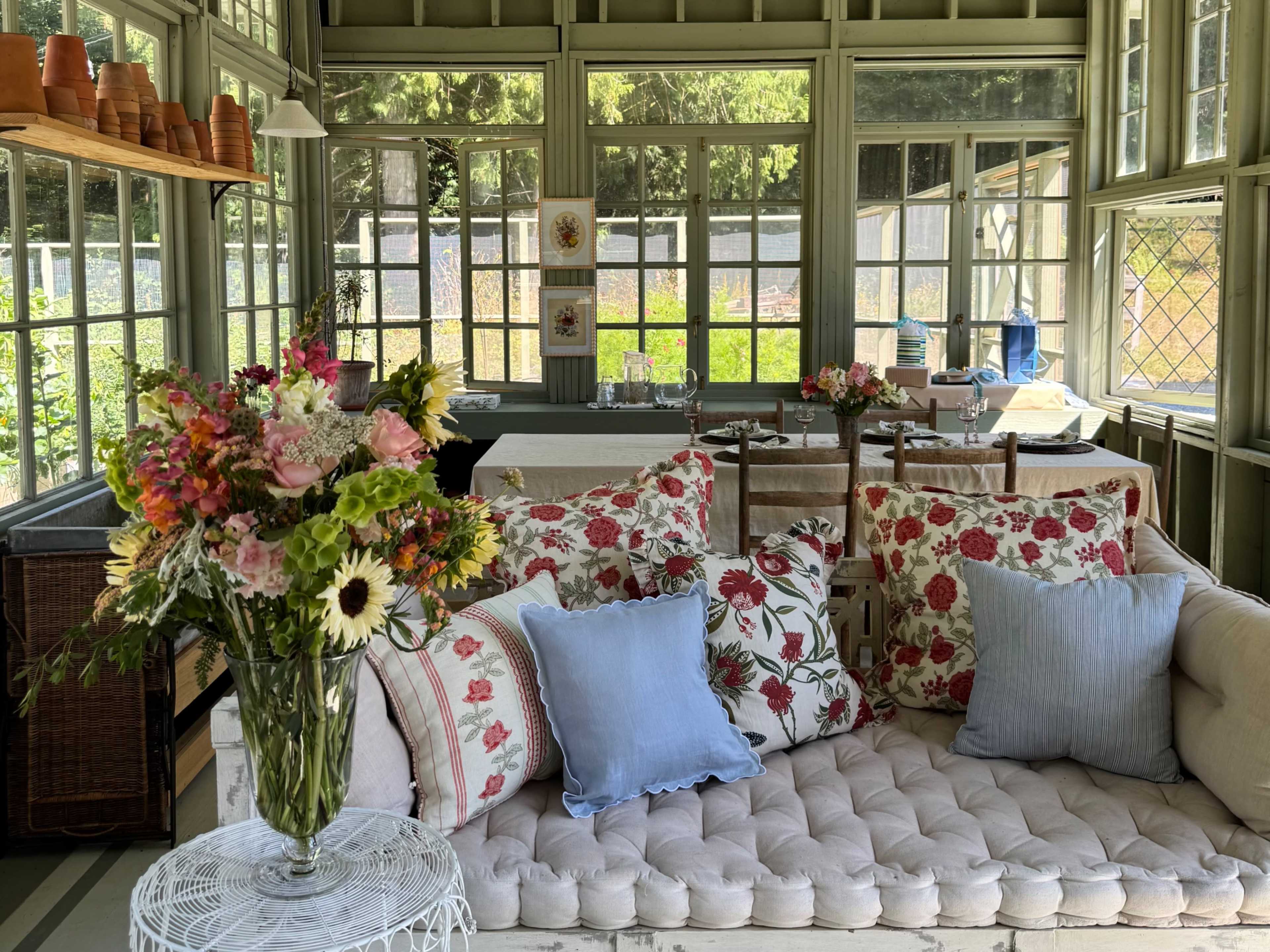 The image shows a sunlit room with large windows, a floral sofa adorned with various patterned cushions, and a vase of flowers on a white table.