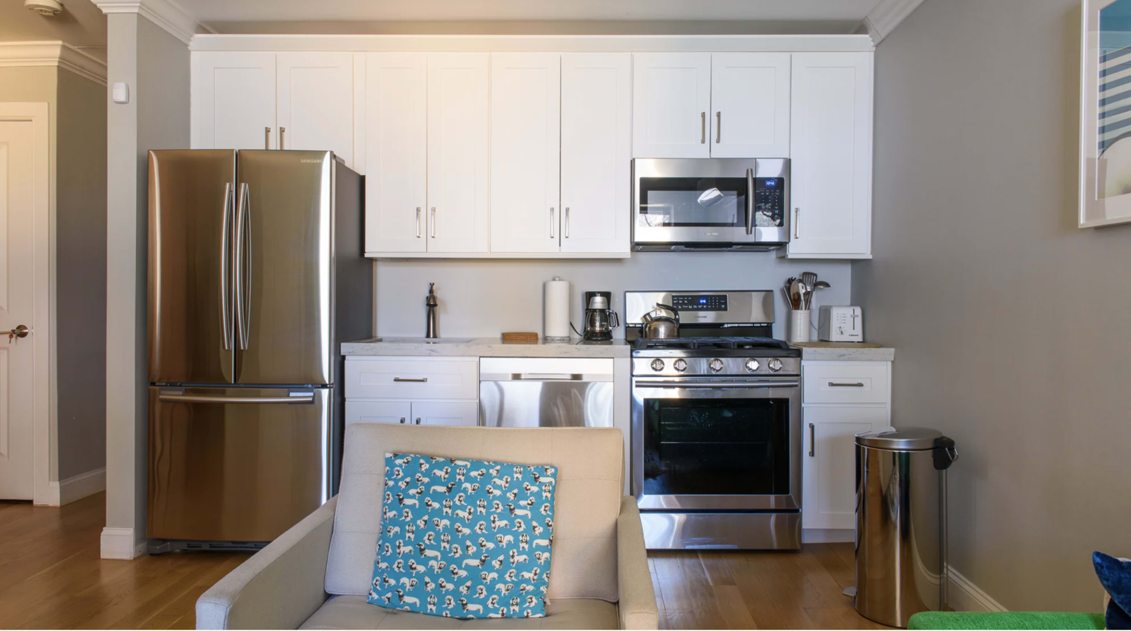 The image shows a modern kitchen with stainless steel appliances, white cabinetry, and a countertop, adjacent to a light-colored living area.