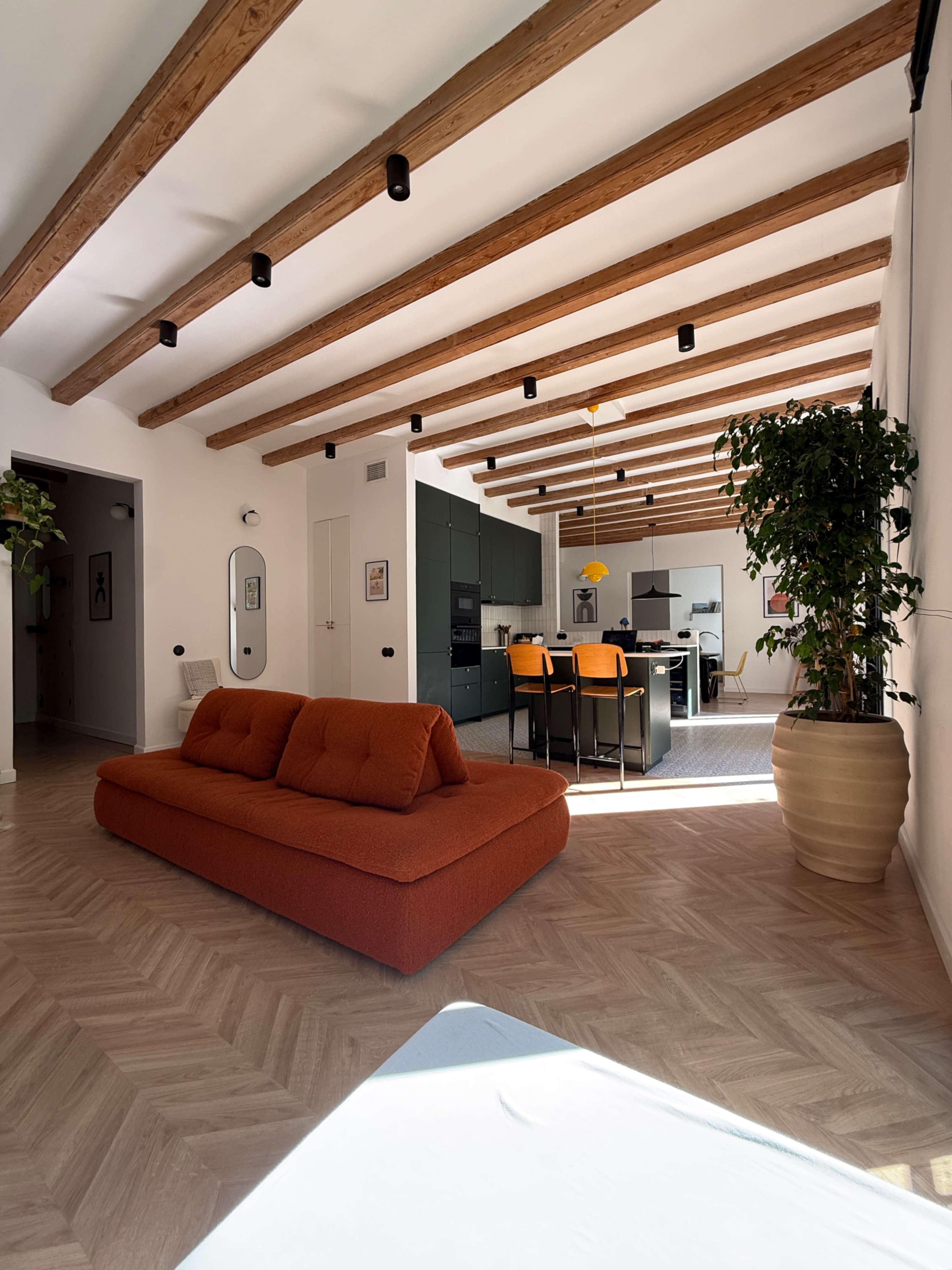 The image shows a bright, modern living room with exposed wooden beams, a large orange sofa, and a view into a kitchen area featuring green cabinets and a dining table with yellow chairs.