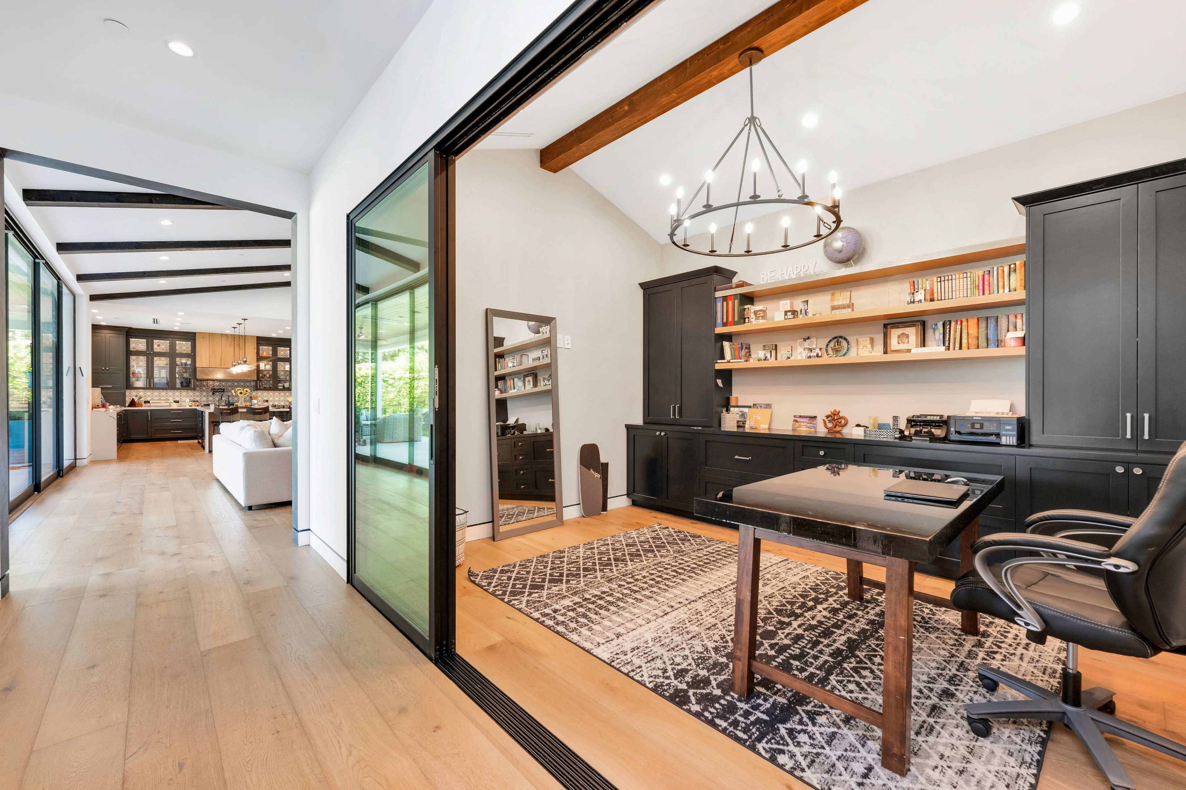 A spacious home office features a large black desk and bookshelves against a light-colored wall, with a view into an adjacent living area through glass doors.