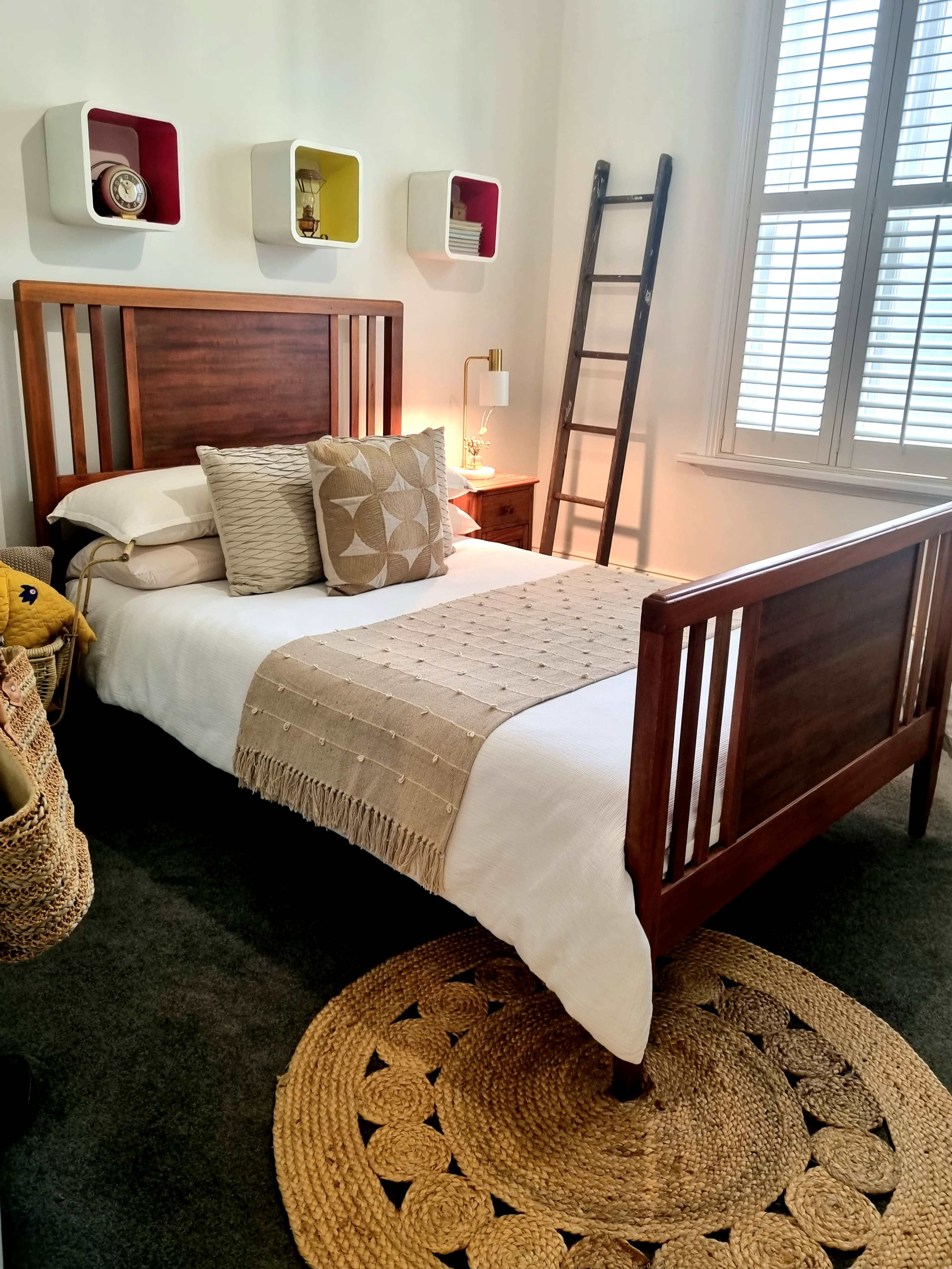 The image shows a neatly arranged bedroom featuring a wooden bed frame with decorative pillows, a textured throw blanket, a ladder against the wall, and colorful wall shelves.