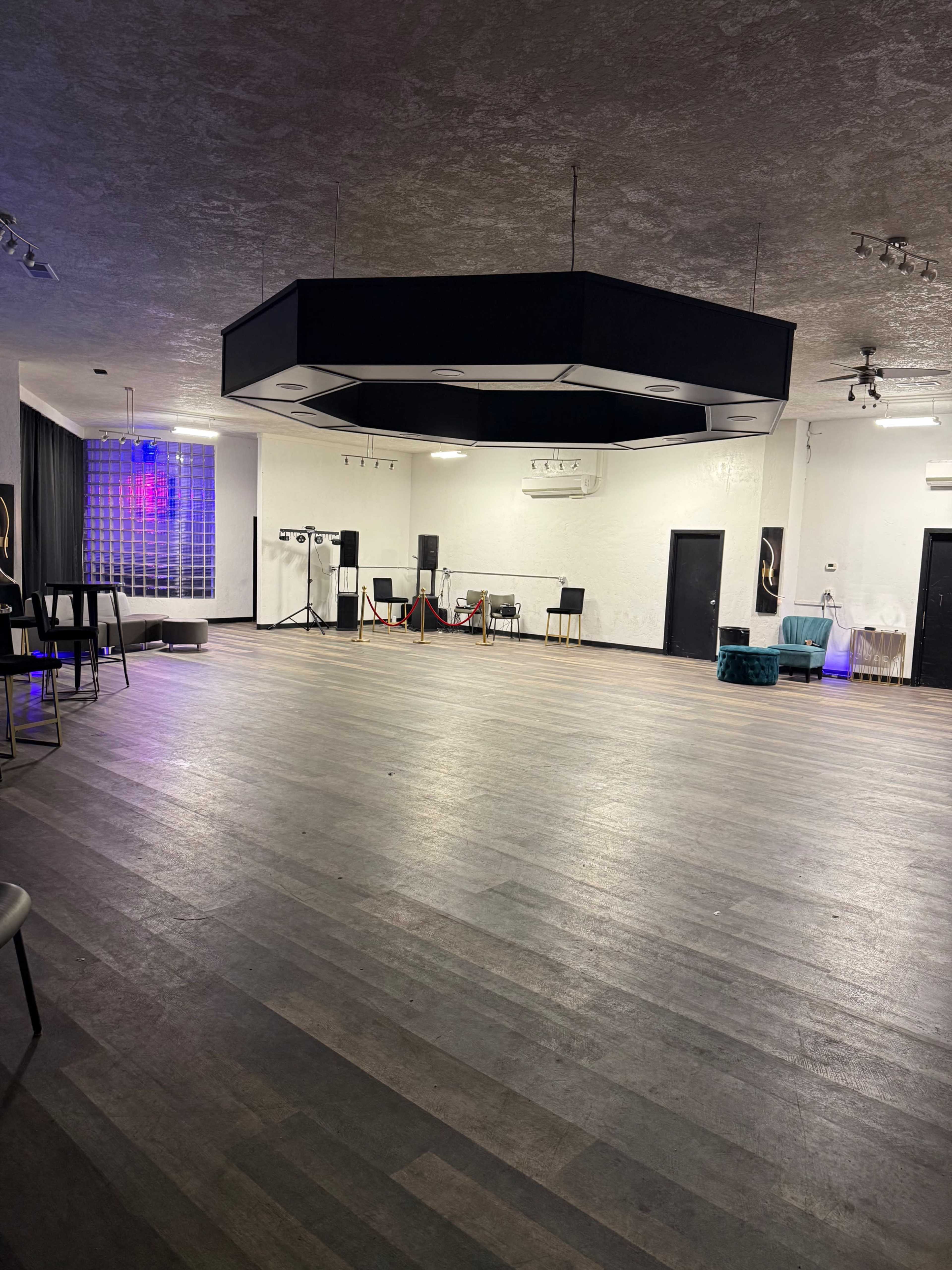 The image shows a spacious, empty event hall with a wooden floor, a central hexagonal ceiling fixture, and various audio equipment set up in the background.