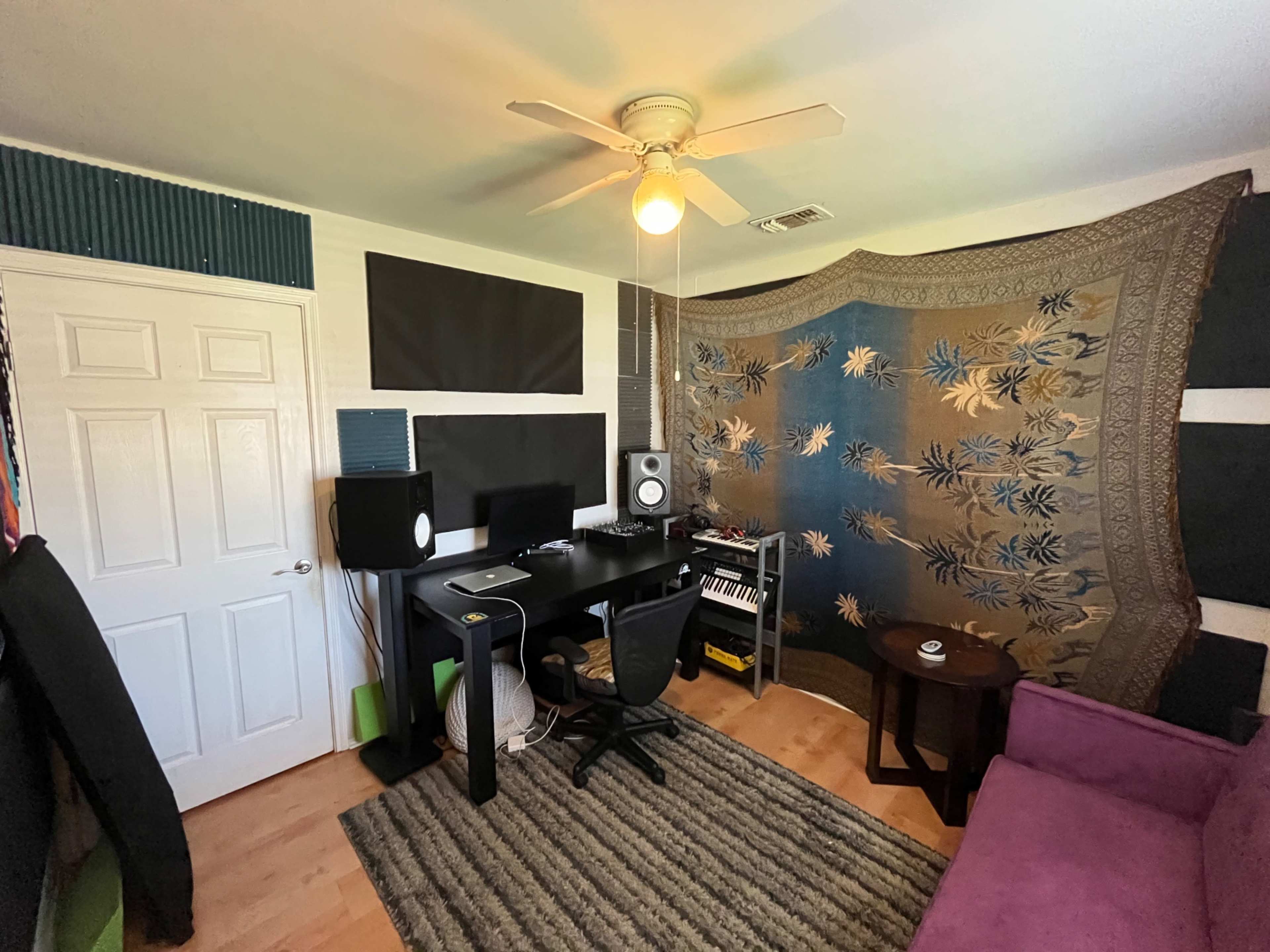 The image shows a small music studio with a desk, speakers, a keyboard, and a patterned wall hanging.