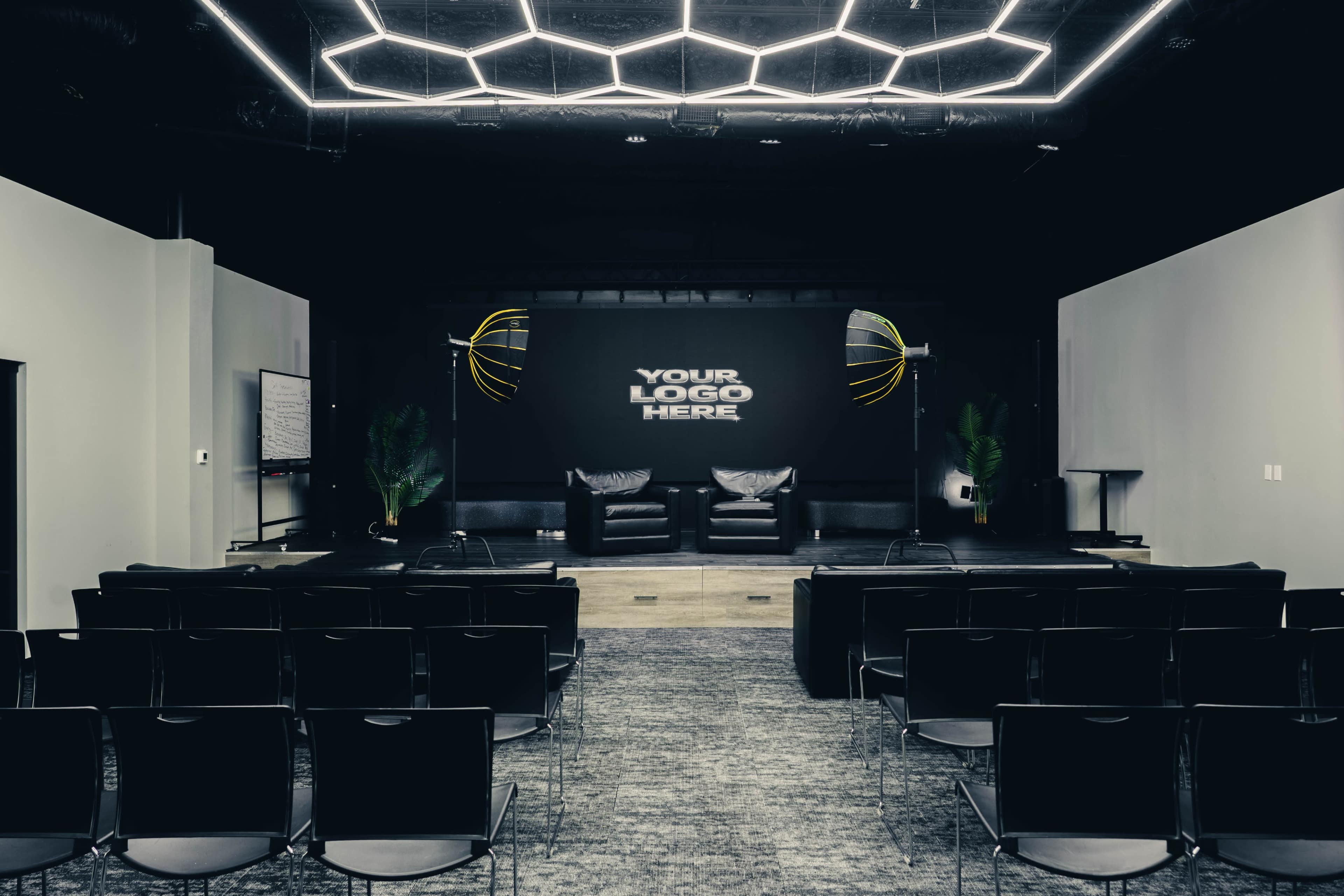 The image depicts a modern event space set up with two black chairs on a stage, surrounded by empty rows of chairs and a large wall displaying a placeholder for a logo.