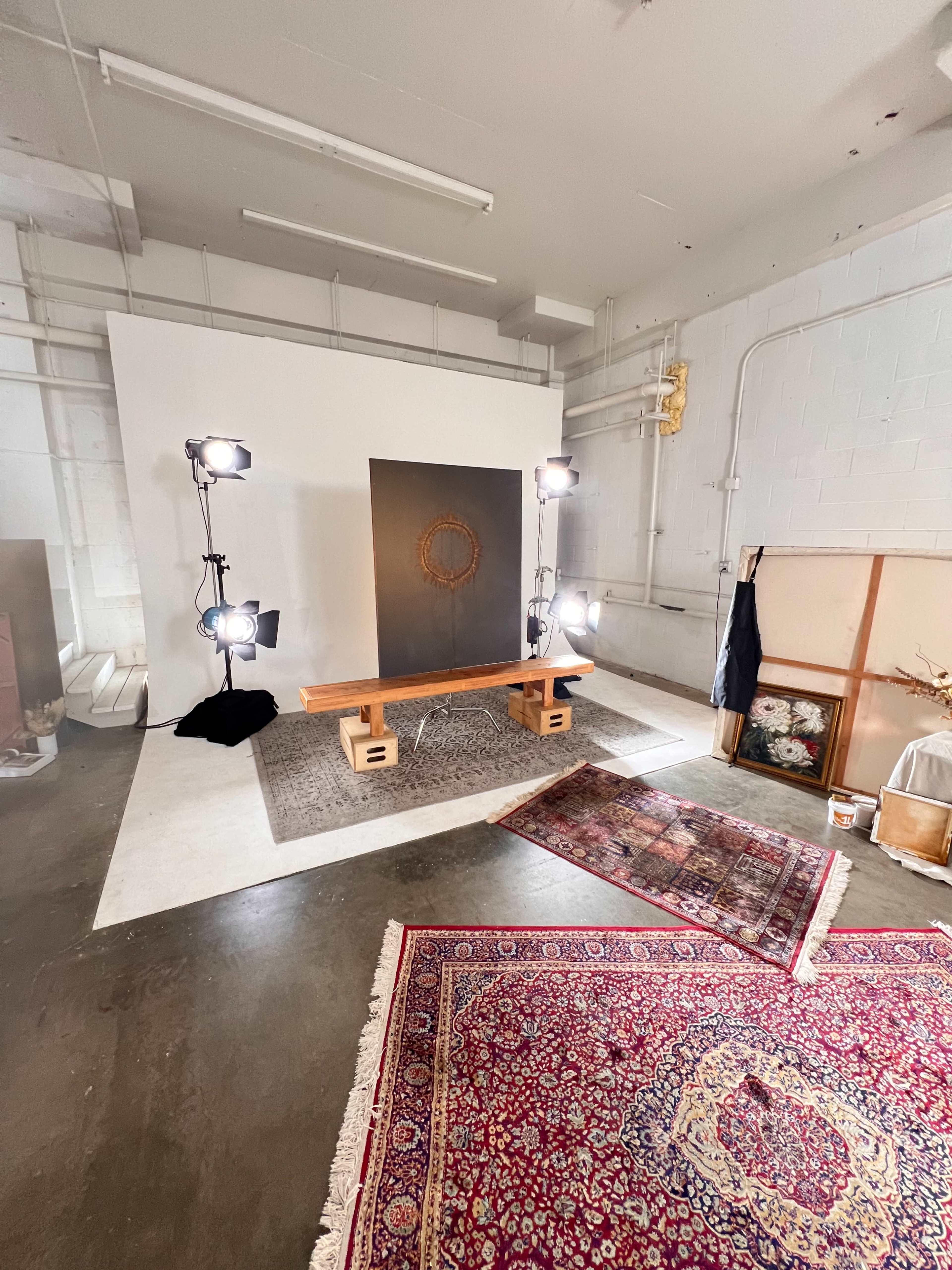 The image shows a photography studio with a wooden bench in front of a large dark backdrop, surrounded by studio lights and an area rug, alongside additional decor elements and props.