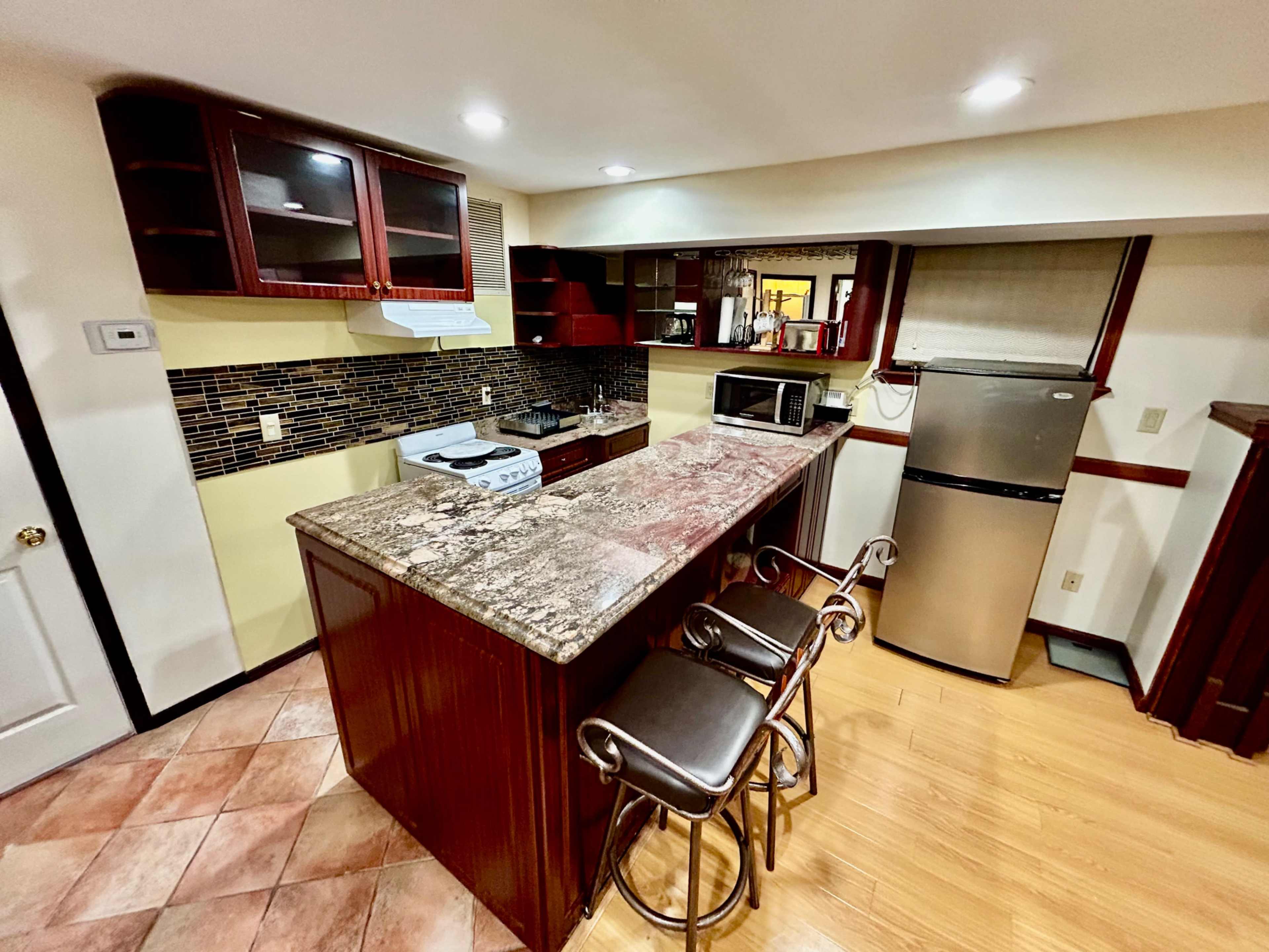 The image shows a modern kitchen with wooden cabinetry, granite countertops, and a breakfast bar with two stools.