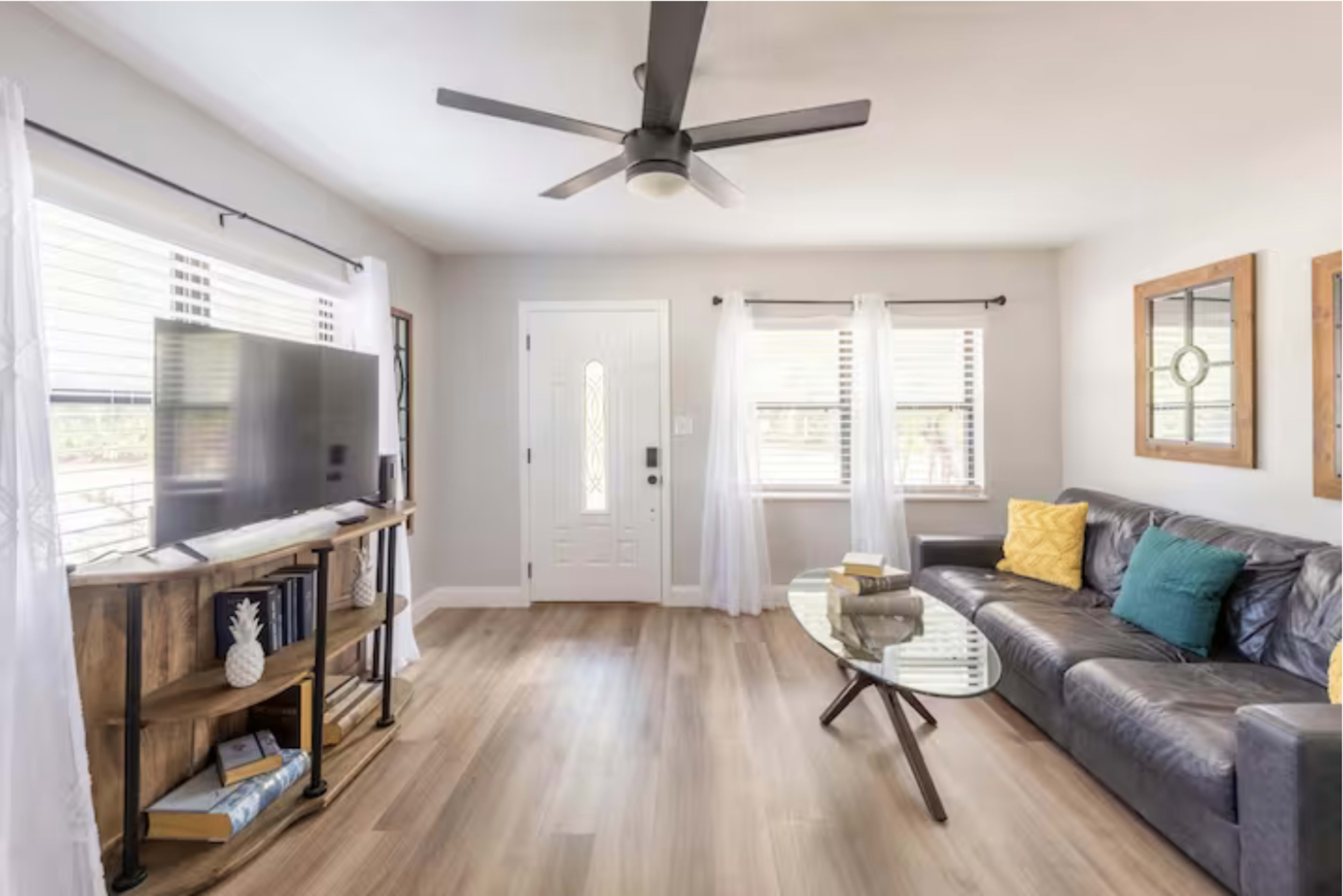The living room features a dark leather sofa, a glass coffee table, a television on a wooden stand, and large windows with sheer white curtains.