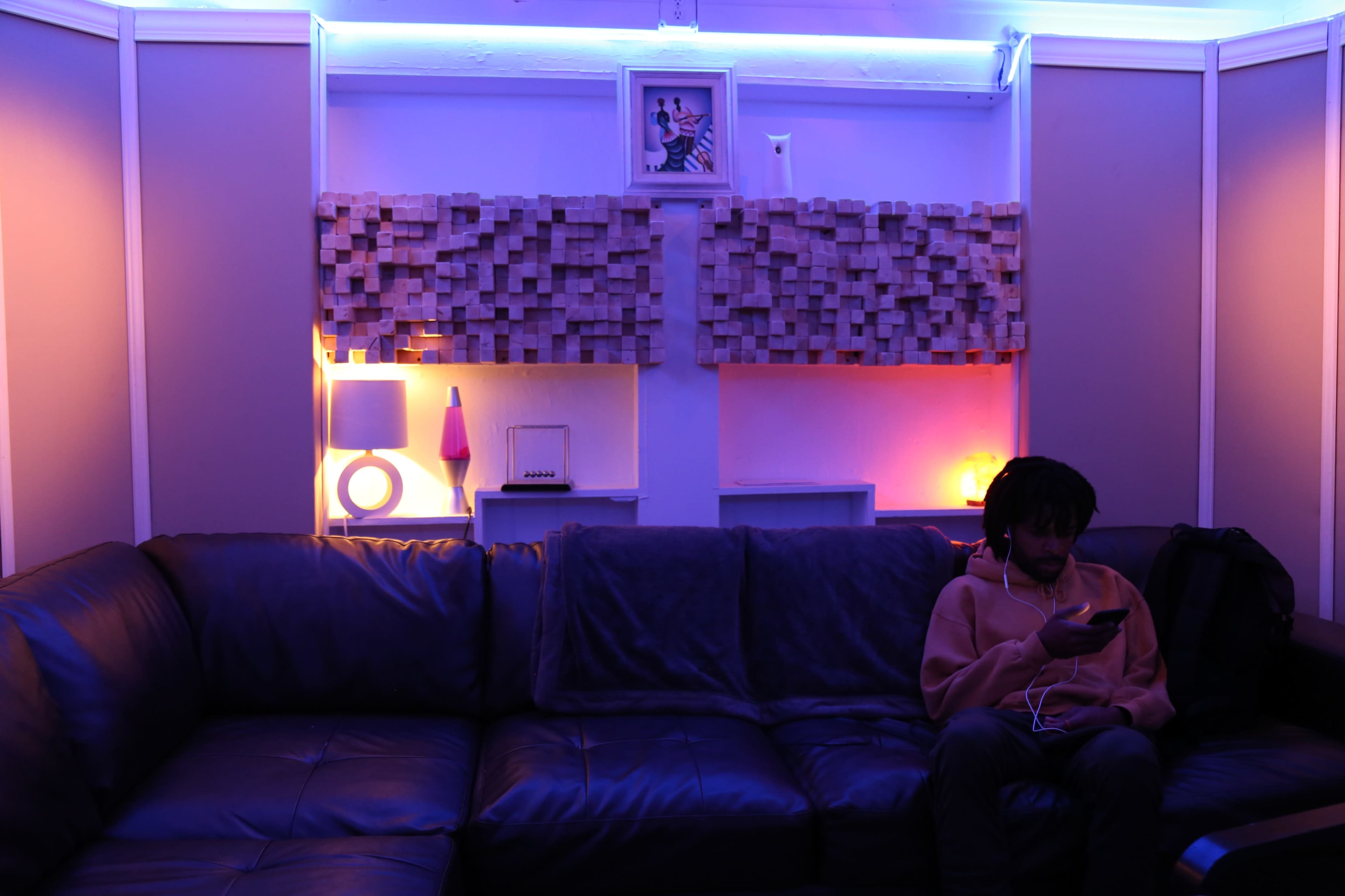 A person sitting on a dark couch is engaged with a smartphone in a softly lit room featuring colorful ambient lighting and decorative wall elements.