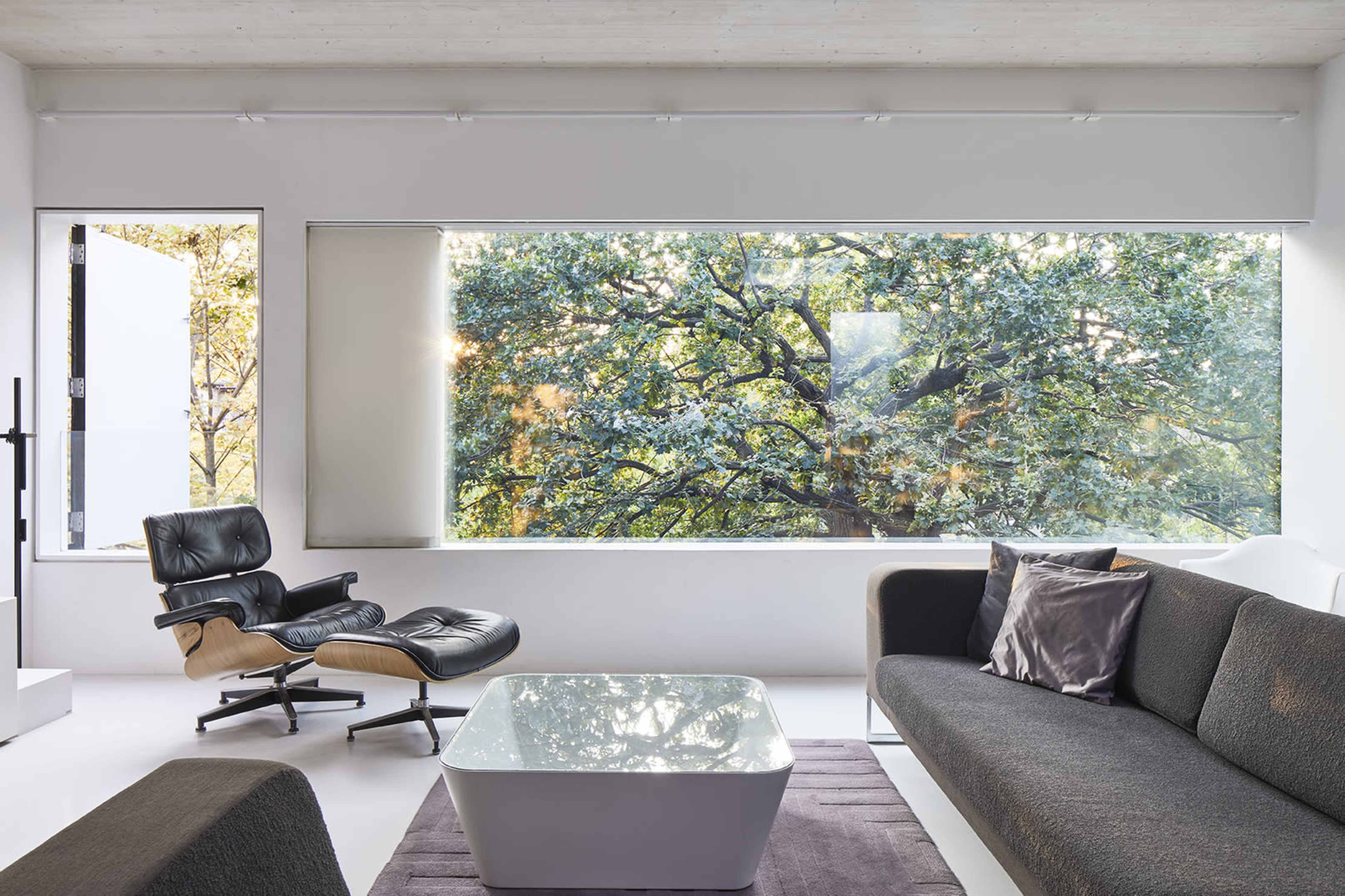 A modern living room featuring a black and beige lounge chair, a white coffee table, and a large window displaying a view of green trees outside.