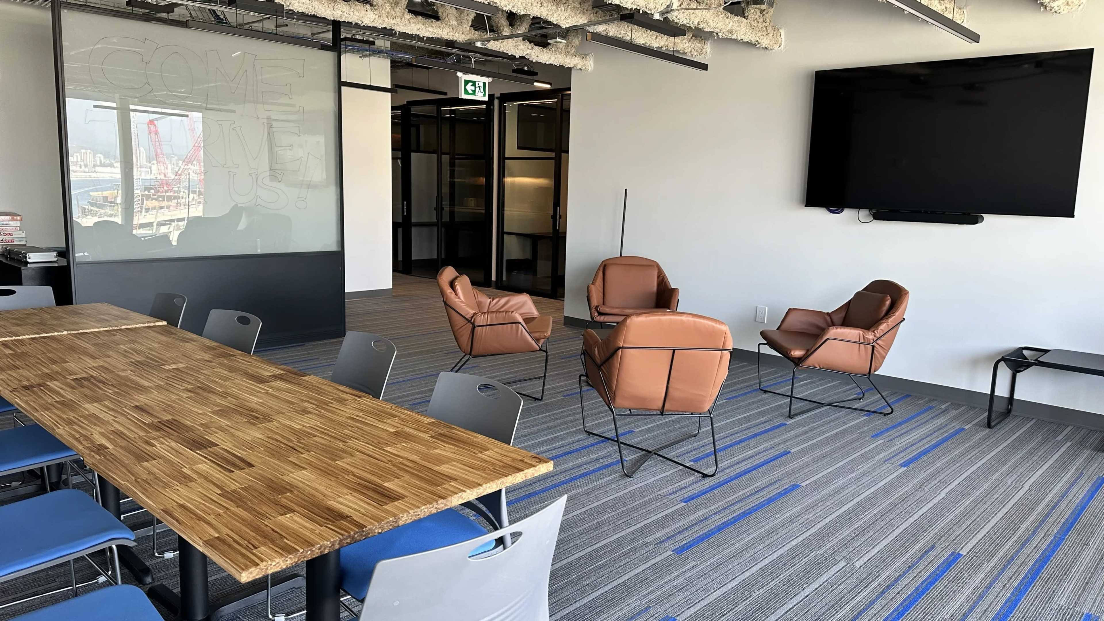 The image shows a modern office space with a long wooden table, several chairs, and a large television mounted on the wall.