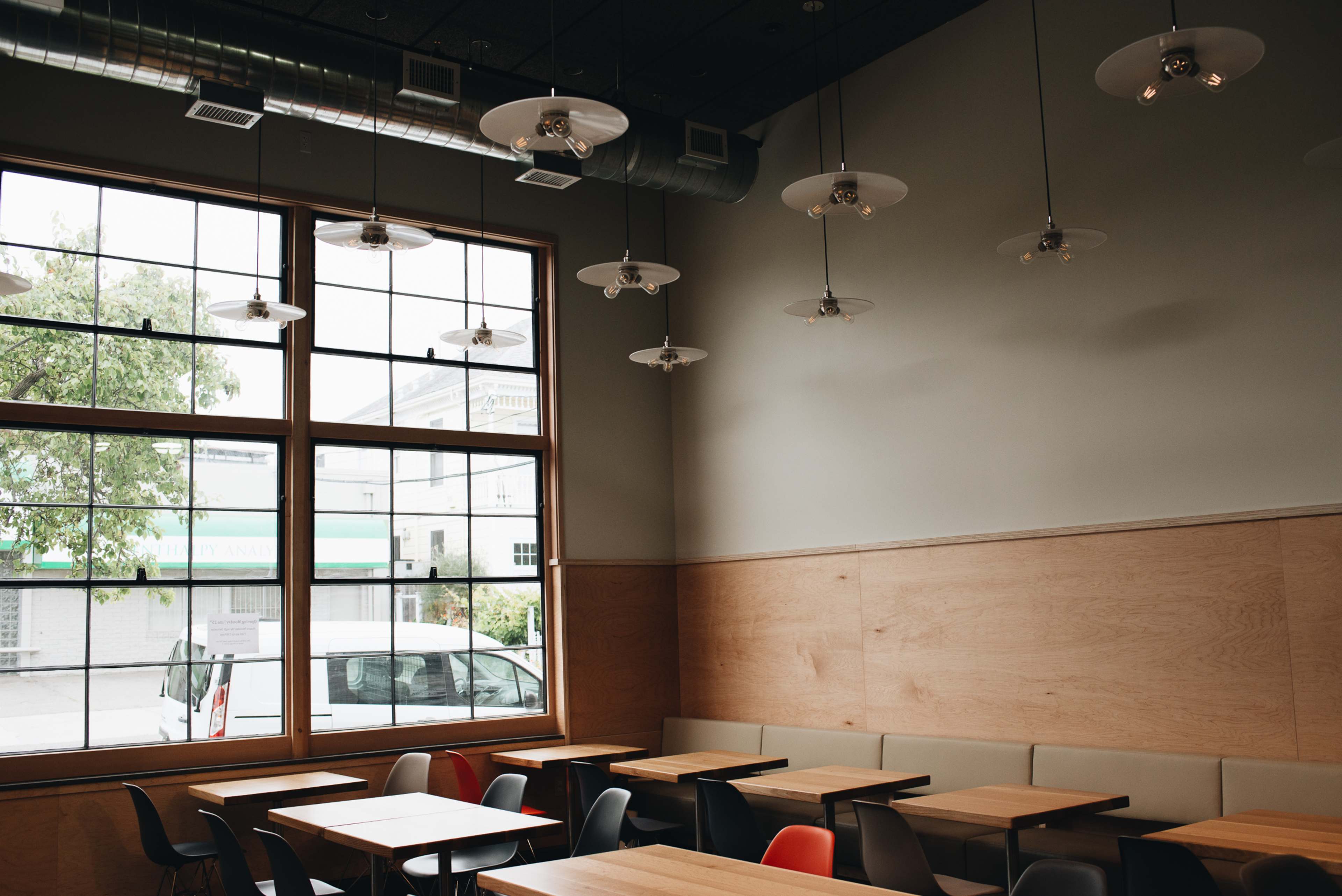 An empty dining area with wooden tables and chairs, large windows letting in natural light, and ceiling fans above.