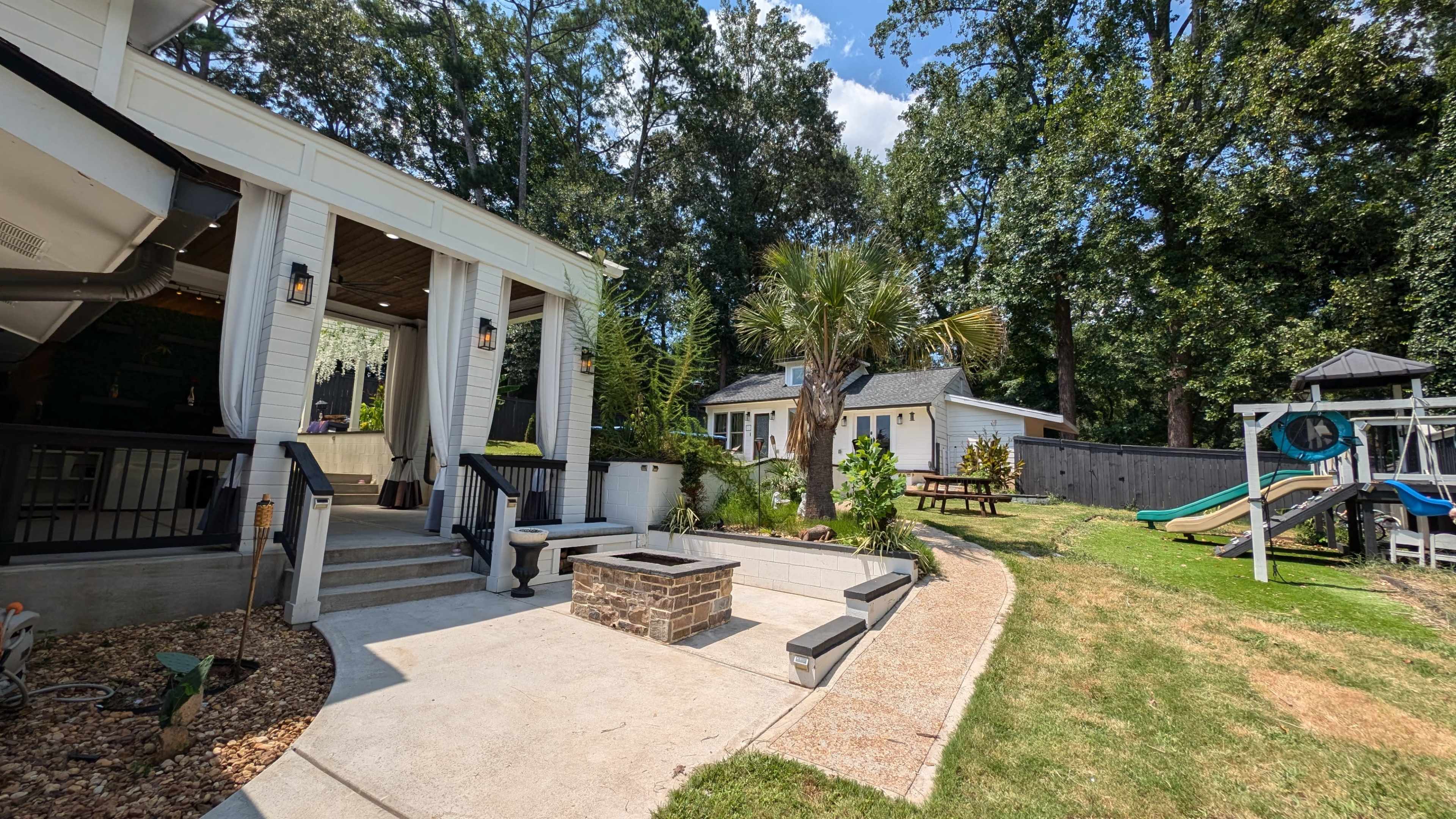 A backyard area with a patio featuring steps and a fire pit, surrounded by a lawn and play equipment, alongside a house and another structure in the background.