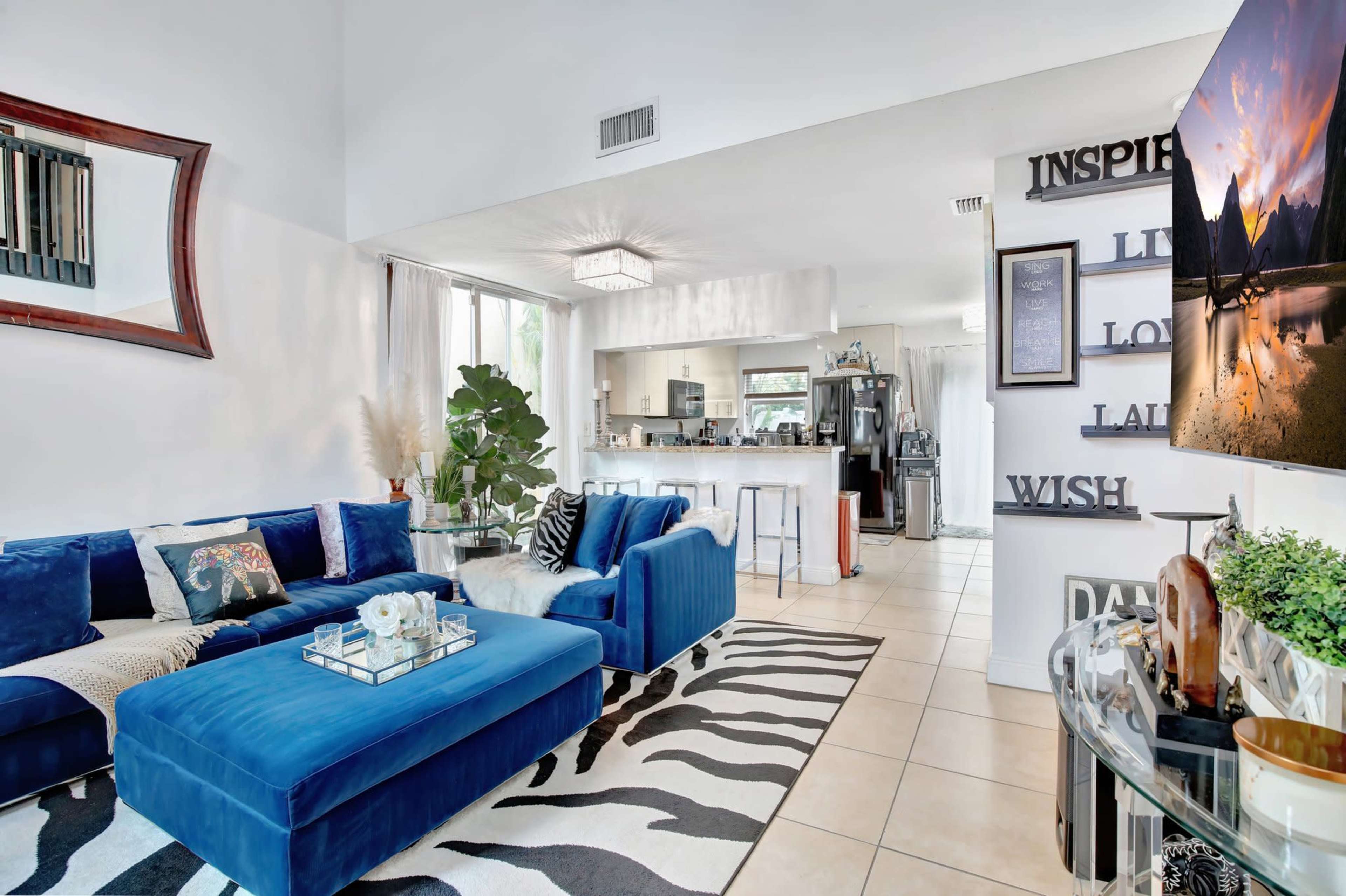 The image shows a bright, modern living room featuring blue seating, a zebra-patterned rug, and an open kitchen area visible in the background.