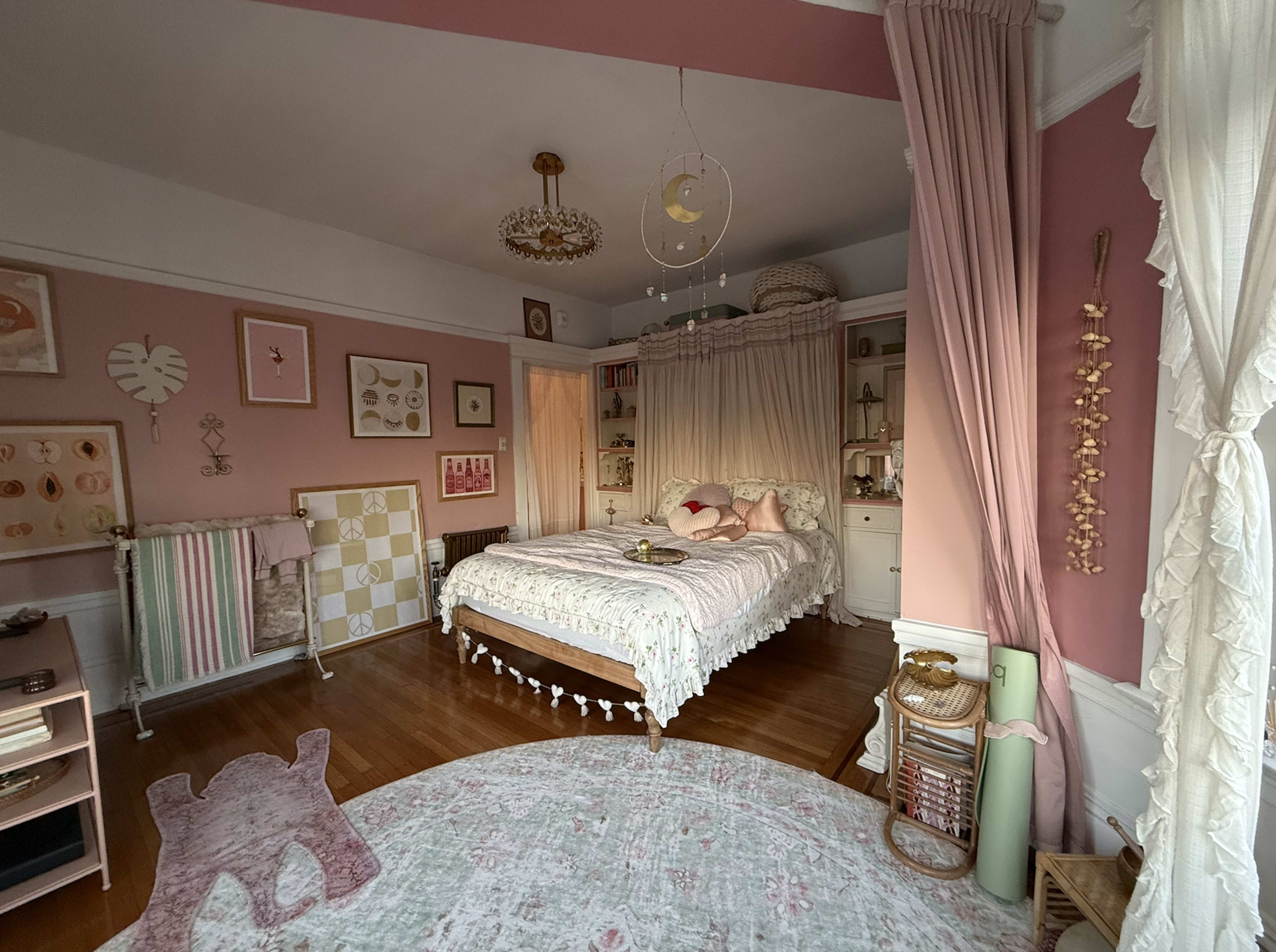 The image shows a softly decorated bedroom featuring a bed with a canopy, pastel pink walls, and various decorative elements including artwork and rugs.