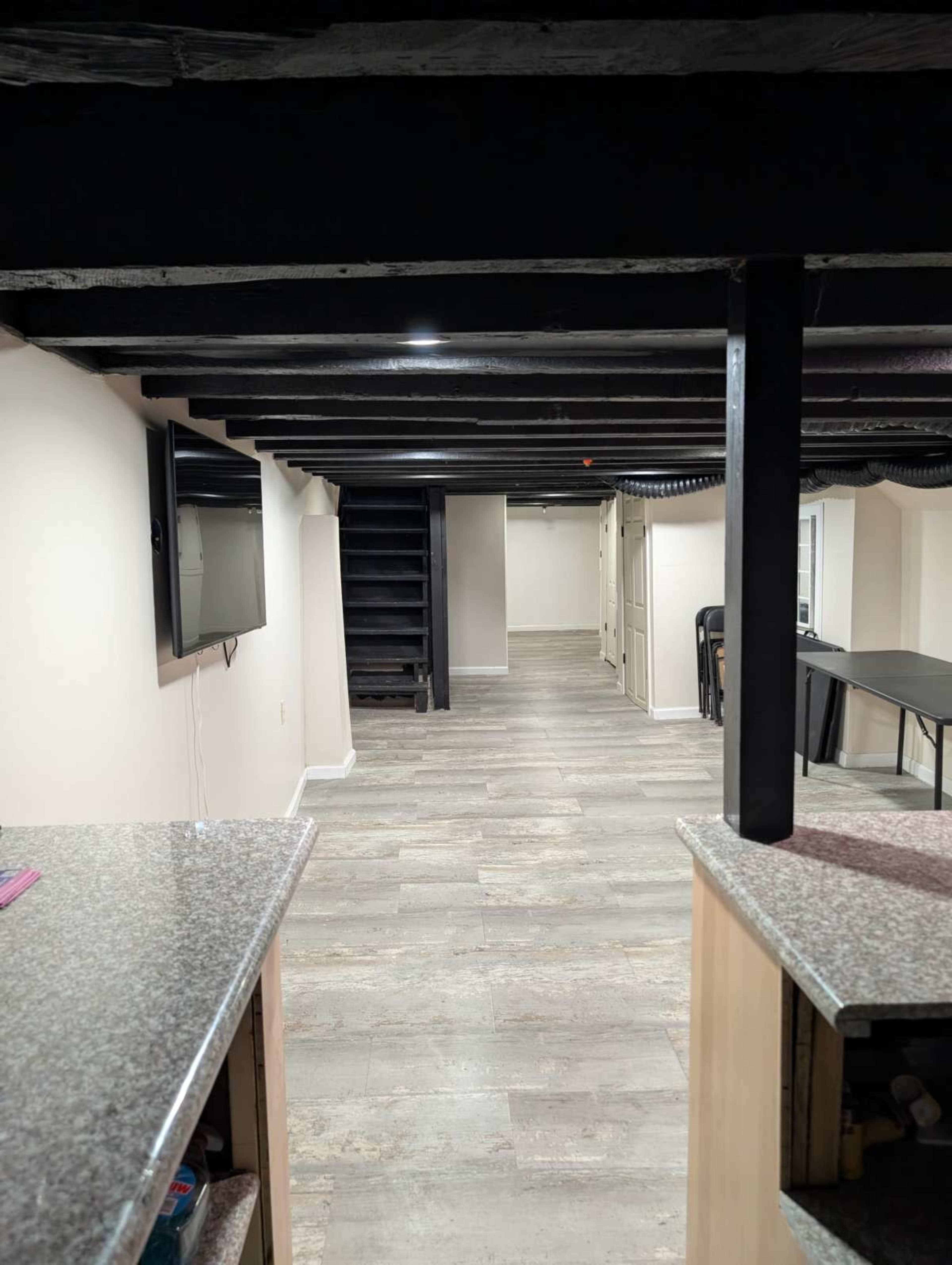 The image shows a finished basement with a polished floor, exposed beams, a television on the left wall, and a staircase leading to the upper level.
