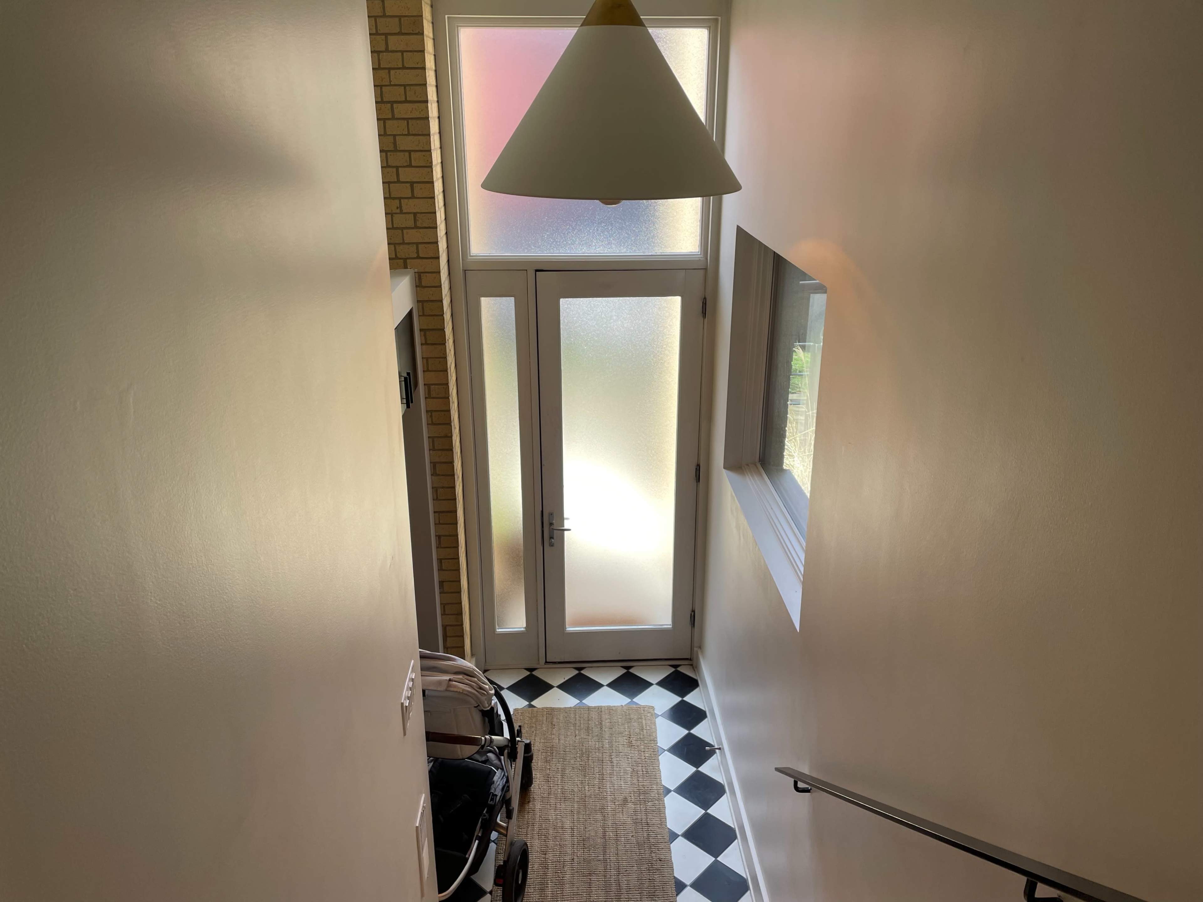 A staircase leads down to a front door with frosted glass panels, adjacent to a large window and a stroller parked on a checkered floor.