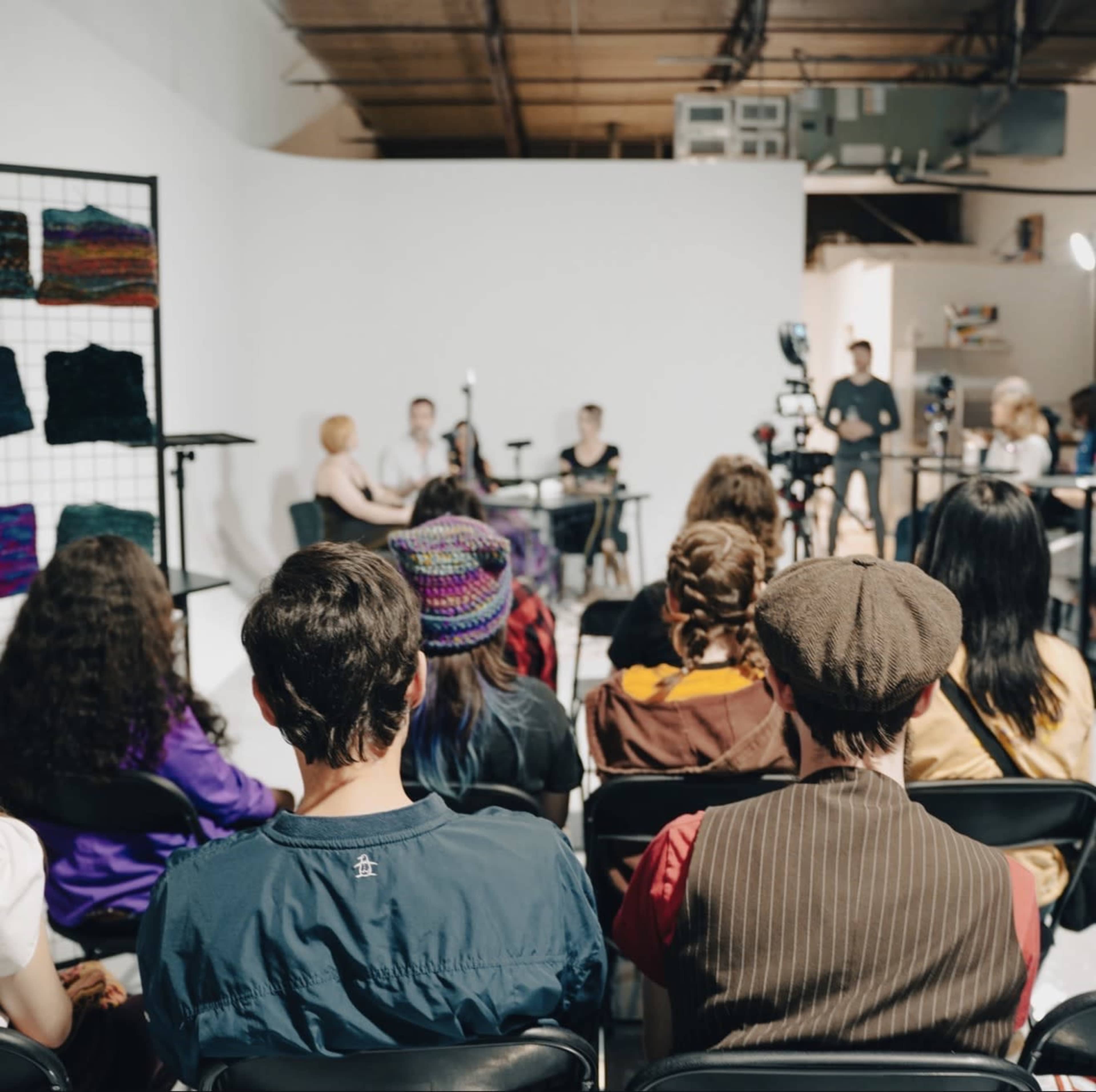 A group of people is seated in a studio, facing a panel discussion taking place at the front, with cameras and lights set up for recording.