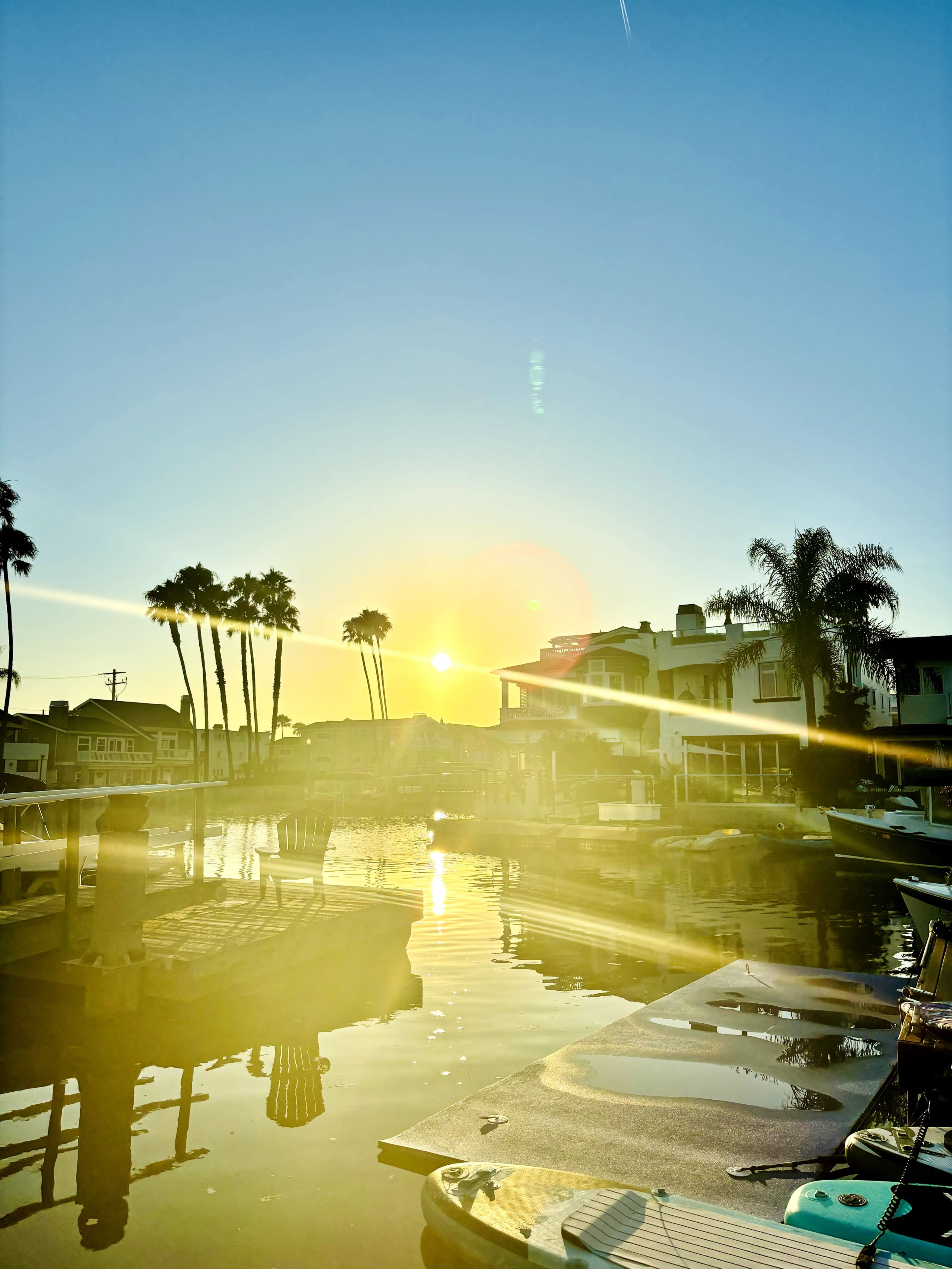 The sun sets over a calm marina, reflecting light on the water while palm trees frame the scene.
