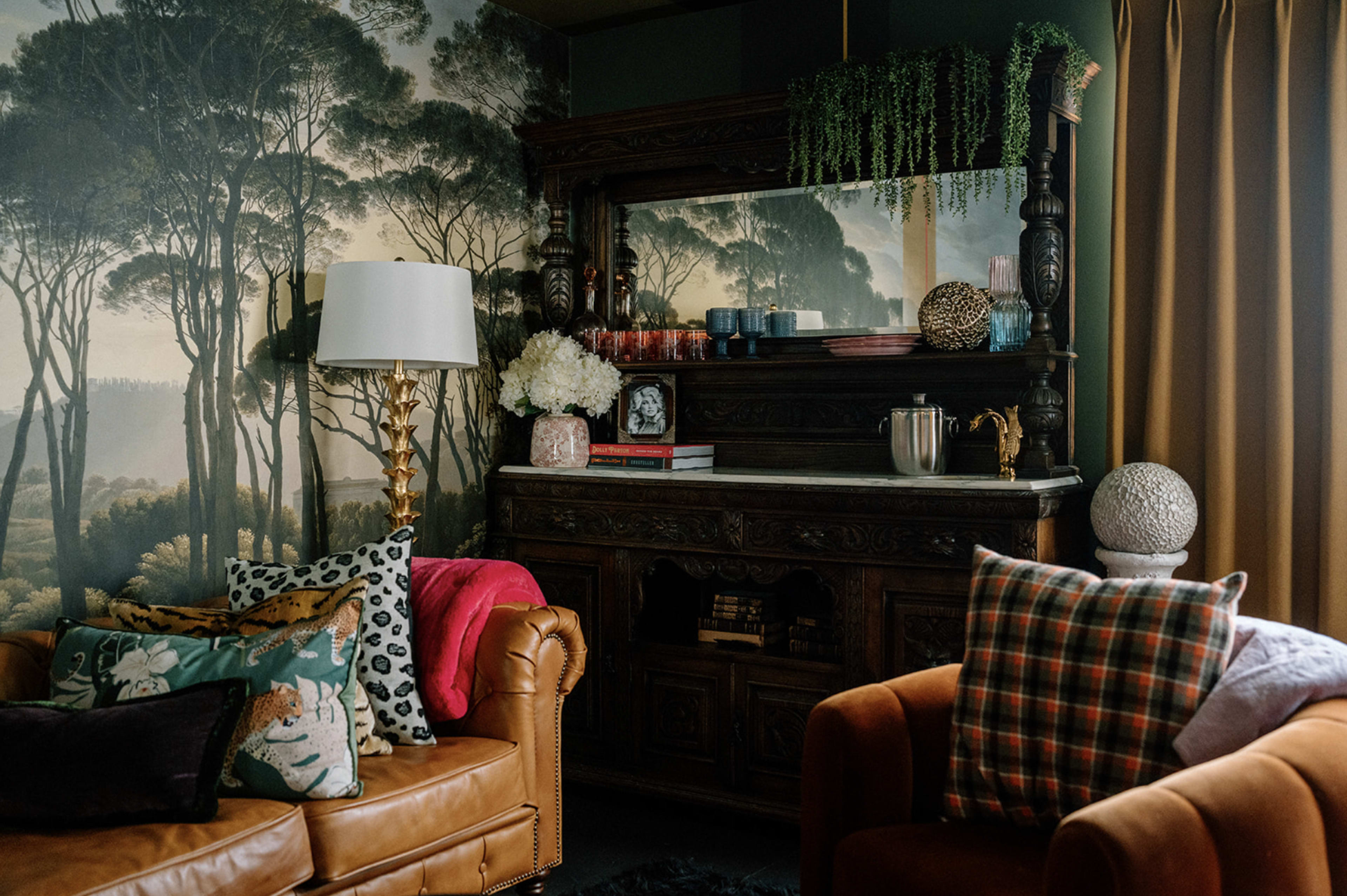 A cozy living room with a vintage wooden sideboard, decorative plants, and a mix of textured cushions on a brown leather couch and an orange armchair.