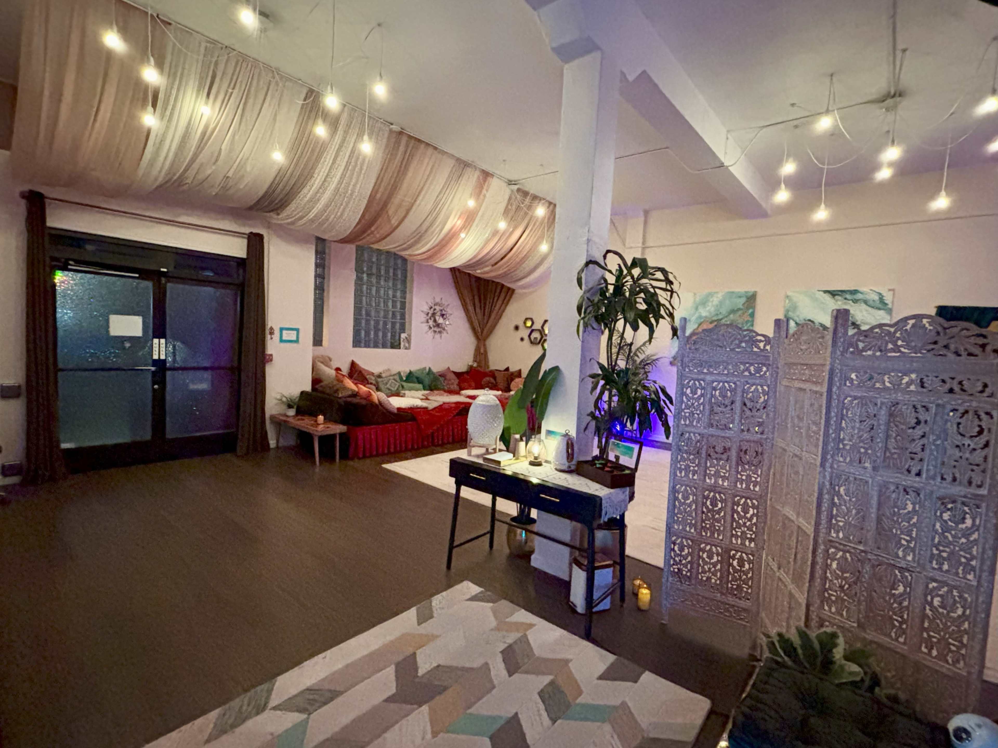 The image shows a cozy indoor space featuring a seating area with cushions, ambient lighting, a decorative partition, and potted plants.