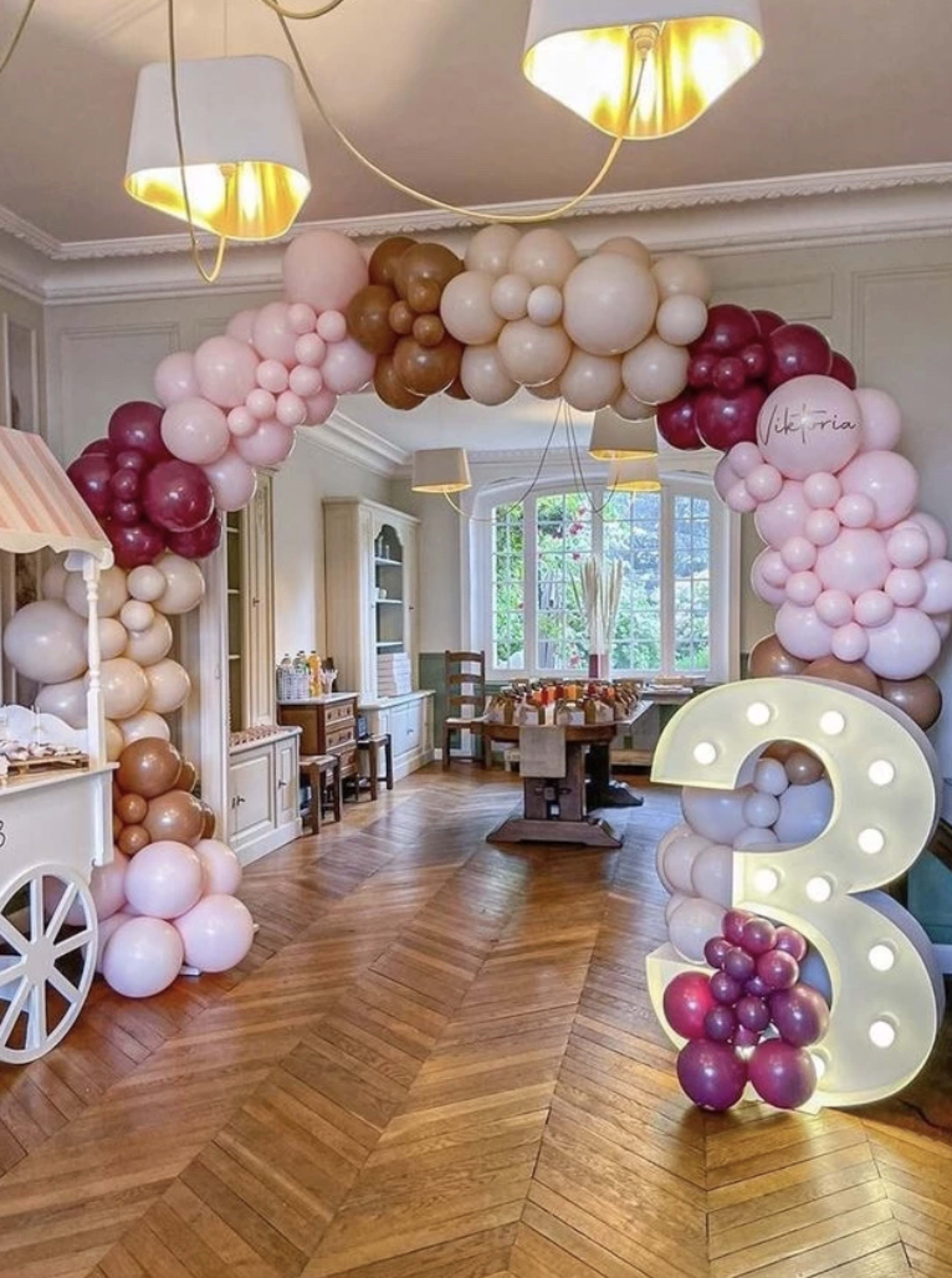 A decorative balloon arch in shades of pink, brown, and burgundy frames a large illuminated number three in a well-lit room with wooden flooring and furniture.