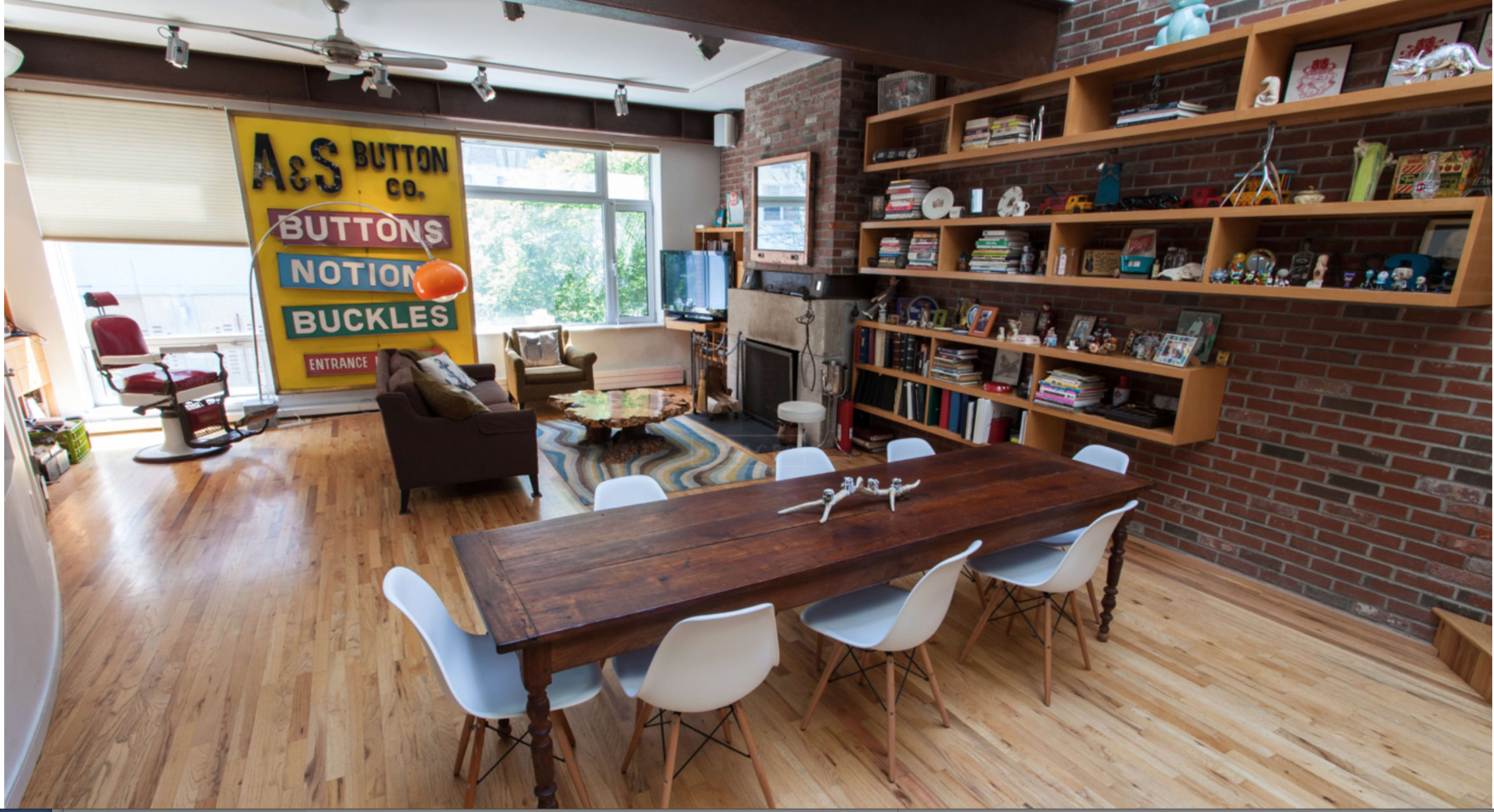 A spacious living area featuring a wooden dining table surrounded by white chairs, with a brick wall and shelves filled with books and decorative items.