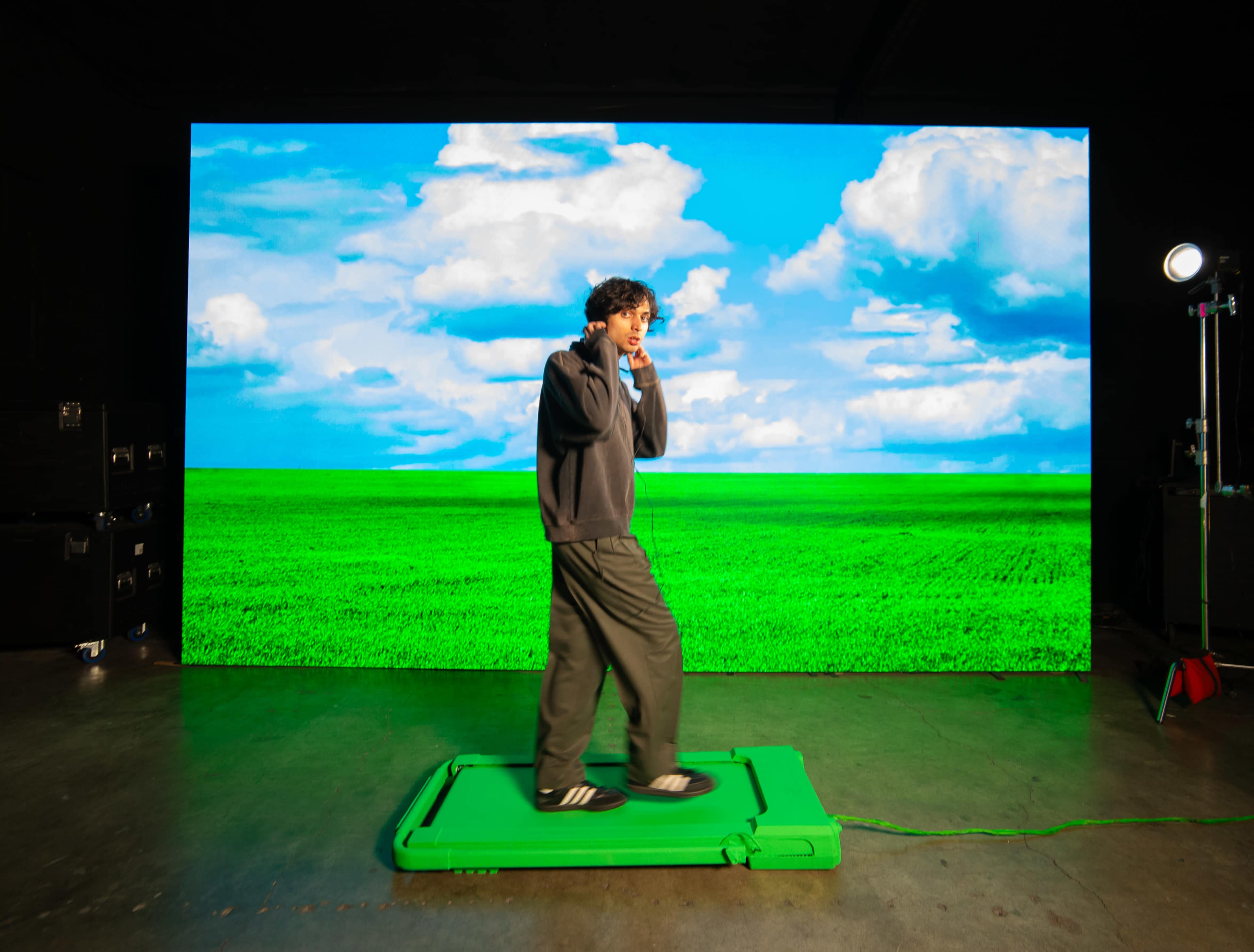 A person stands on a green platform in front of a large screen displaying a blue sky and green field.