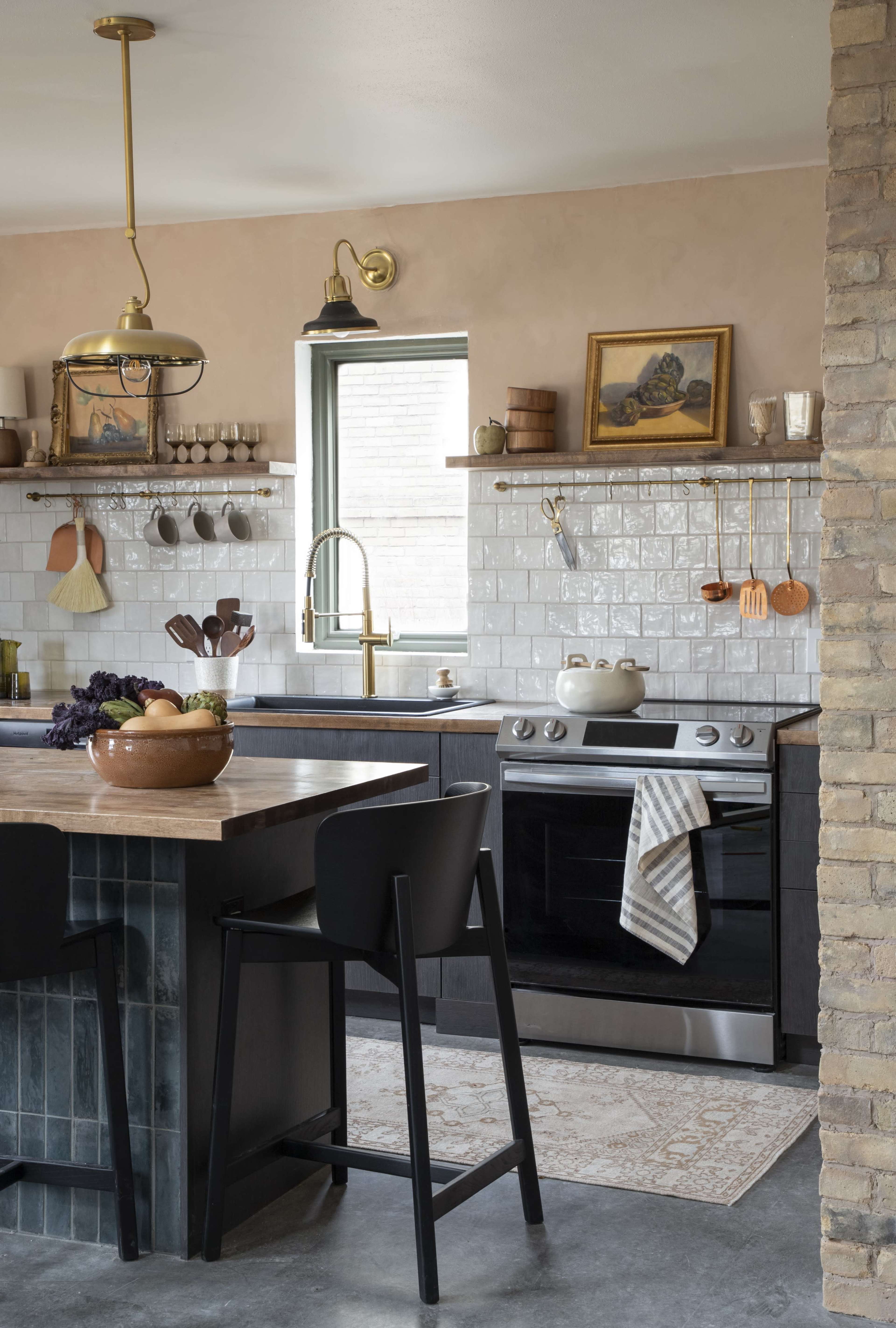 A modern kitchen featuring a central island with bar stools, a black stove, white tiled backsplash, and wooden shelves displaying kitchenware.