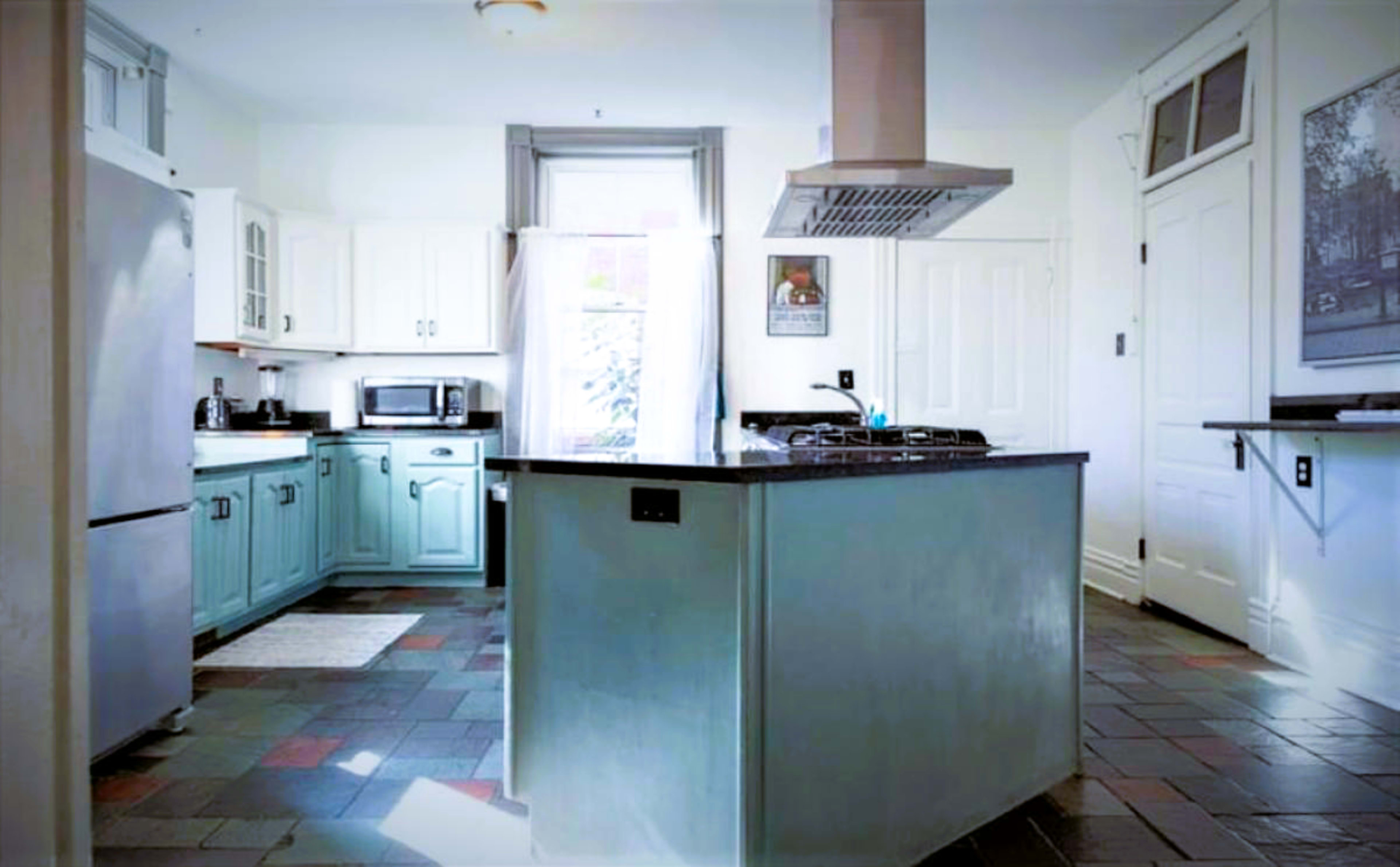 The image shows a modern kitchen with light blue cabinets, a central island with a dark countertop, and a mix of tiled flooring.