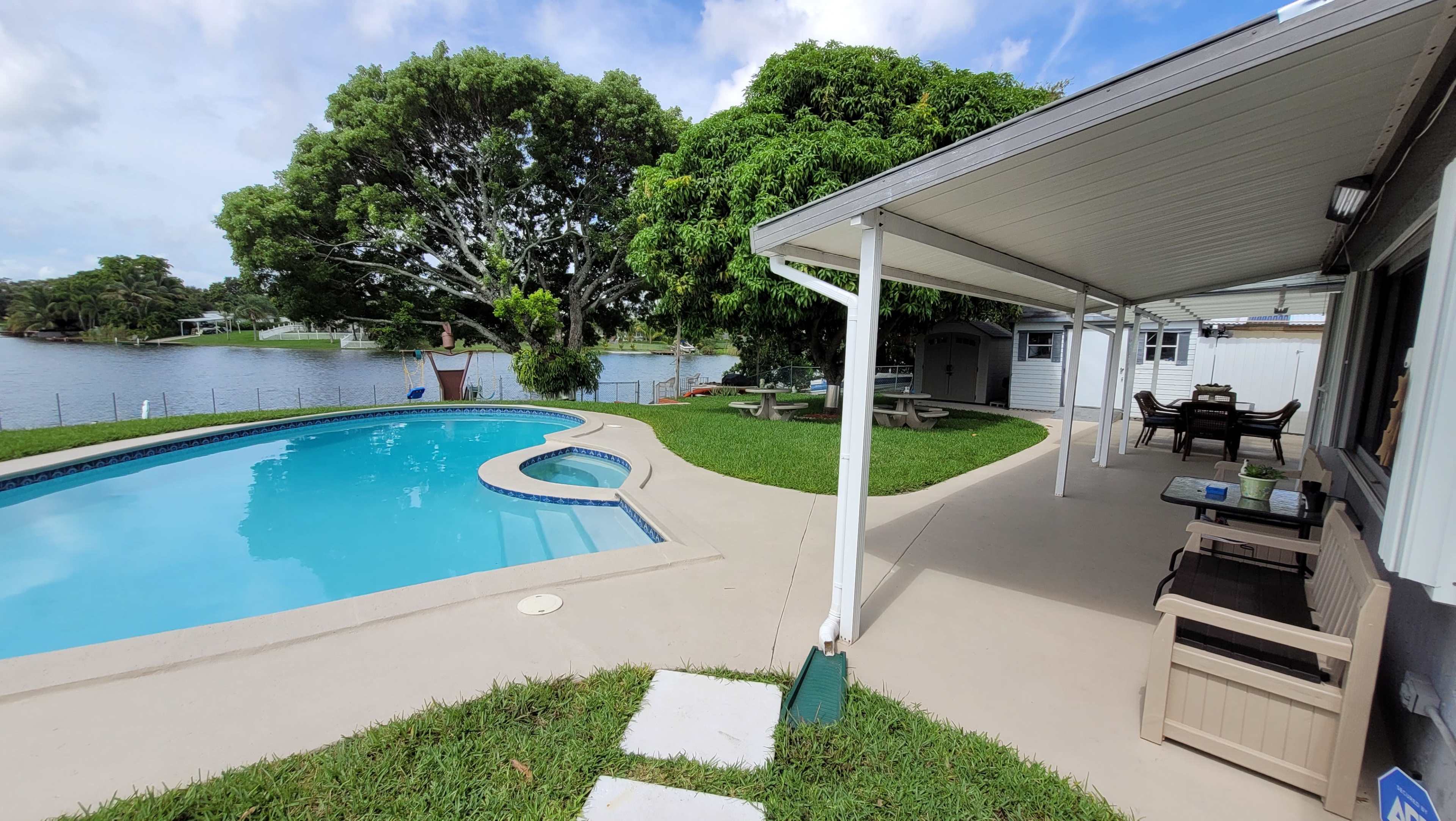 A backyard with a swimming pool, a covered patio area, and a view of a lake surrounded by trees.