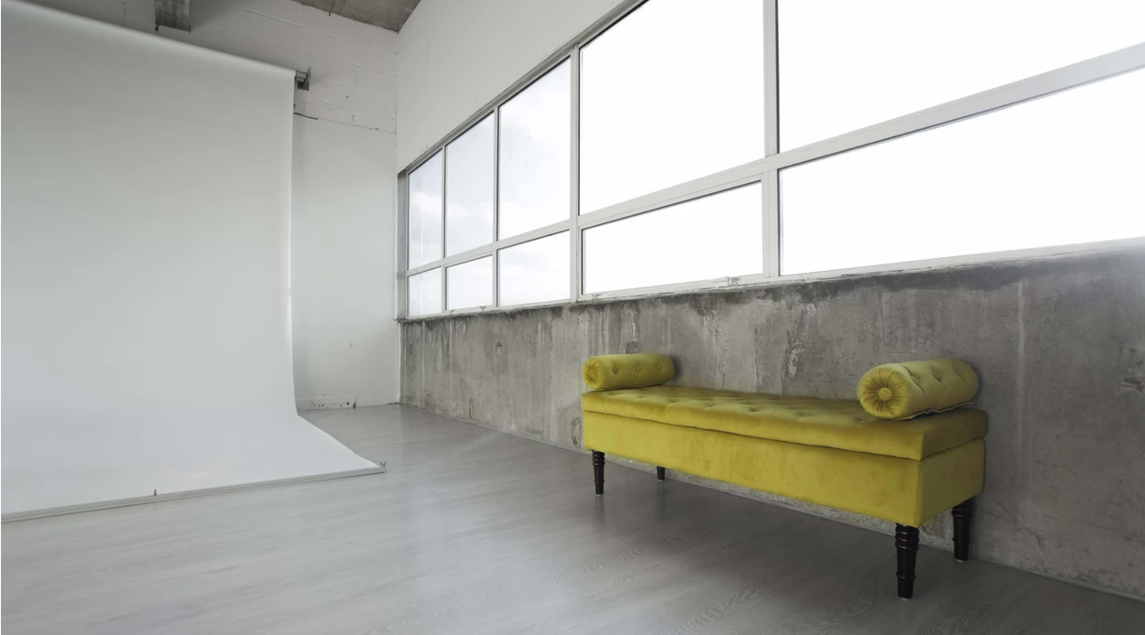 A minimalist room with a yellow upholstered bench beside large windows and a white backdrop.