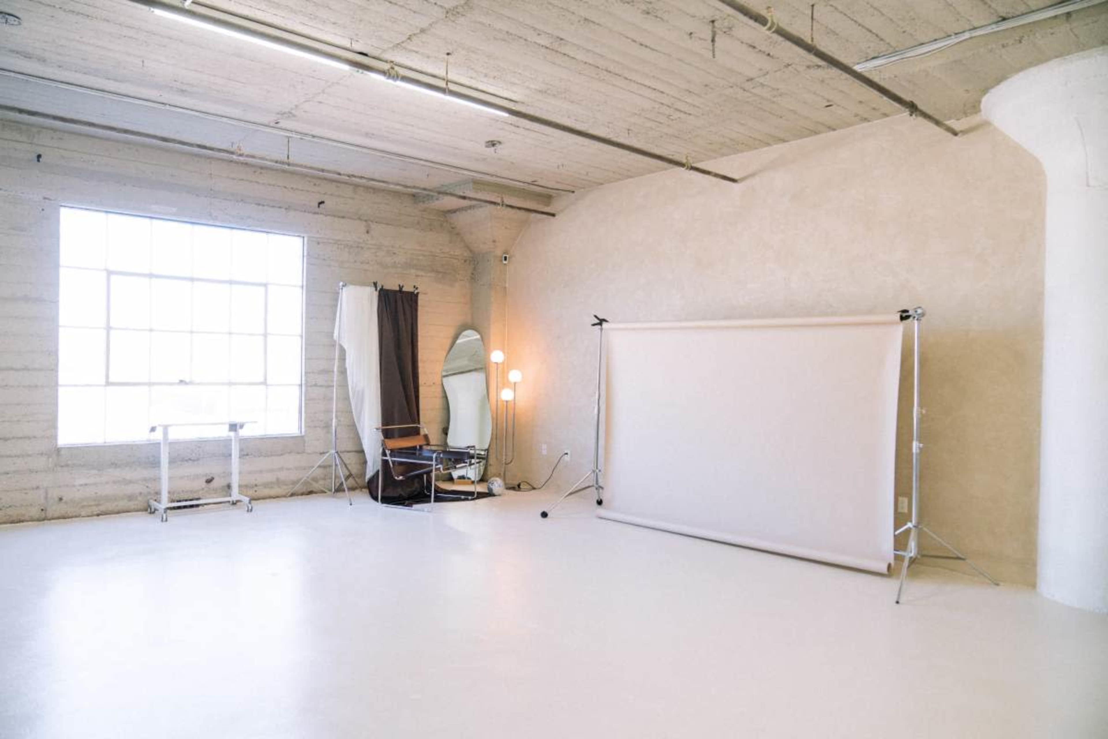 The image shows a spacious photography studio with a large window, a backdrop on stands, a mirror, and a chair.
