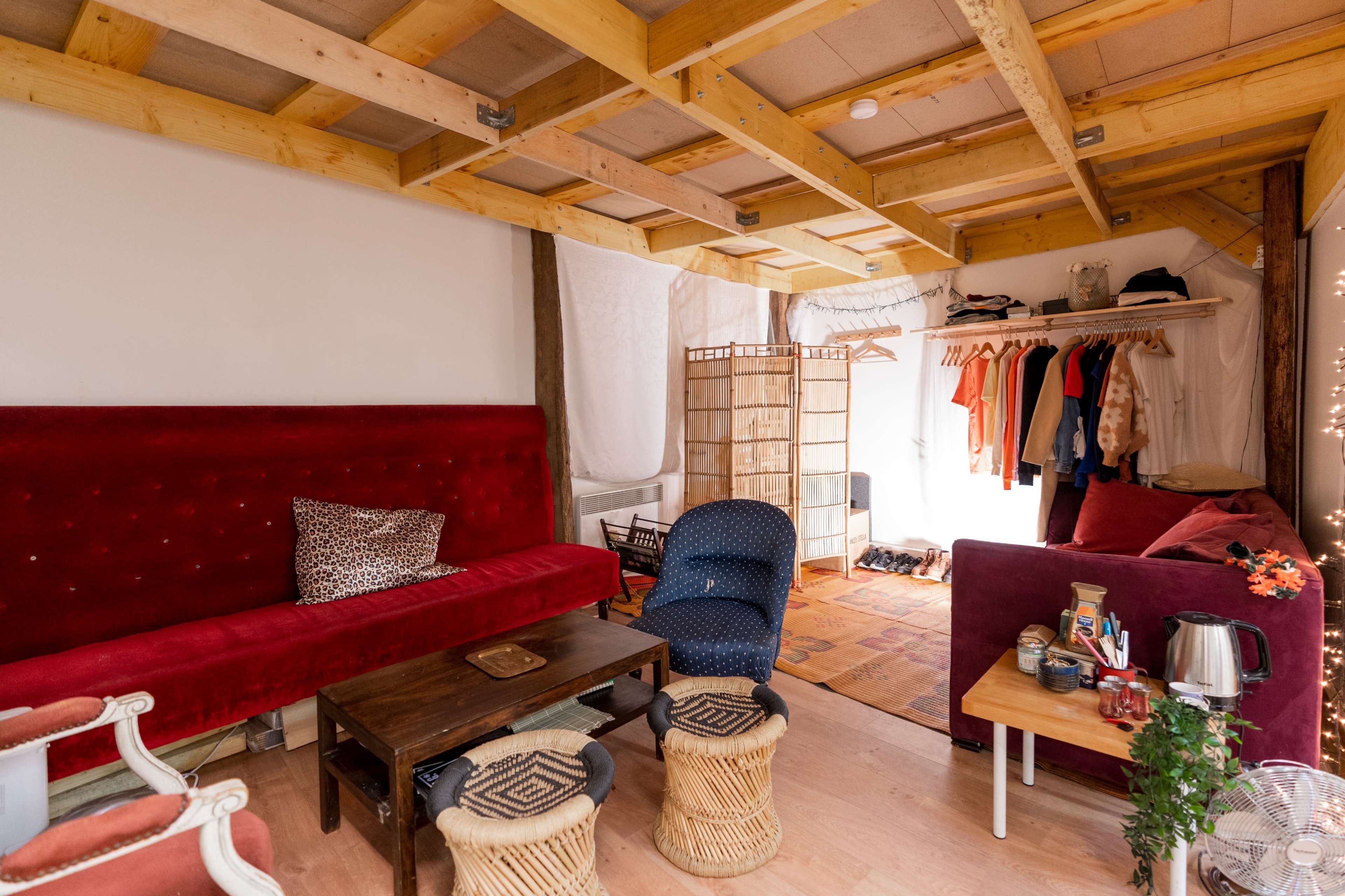 A cozy interior space with a red velvet sofa, a wooden table, and a clothing rack, complemented by natural wood beams and light-colored walls.