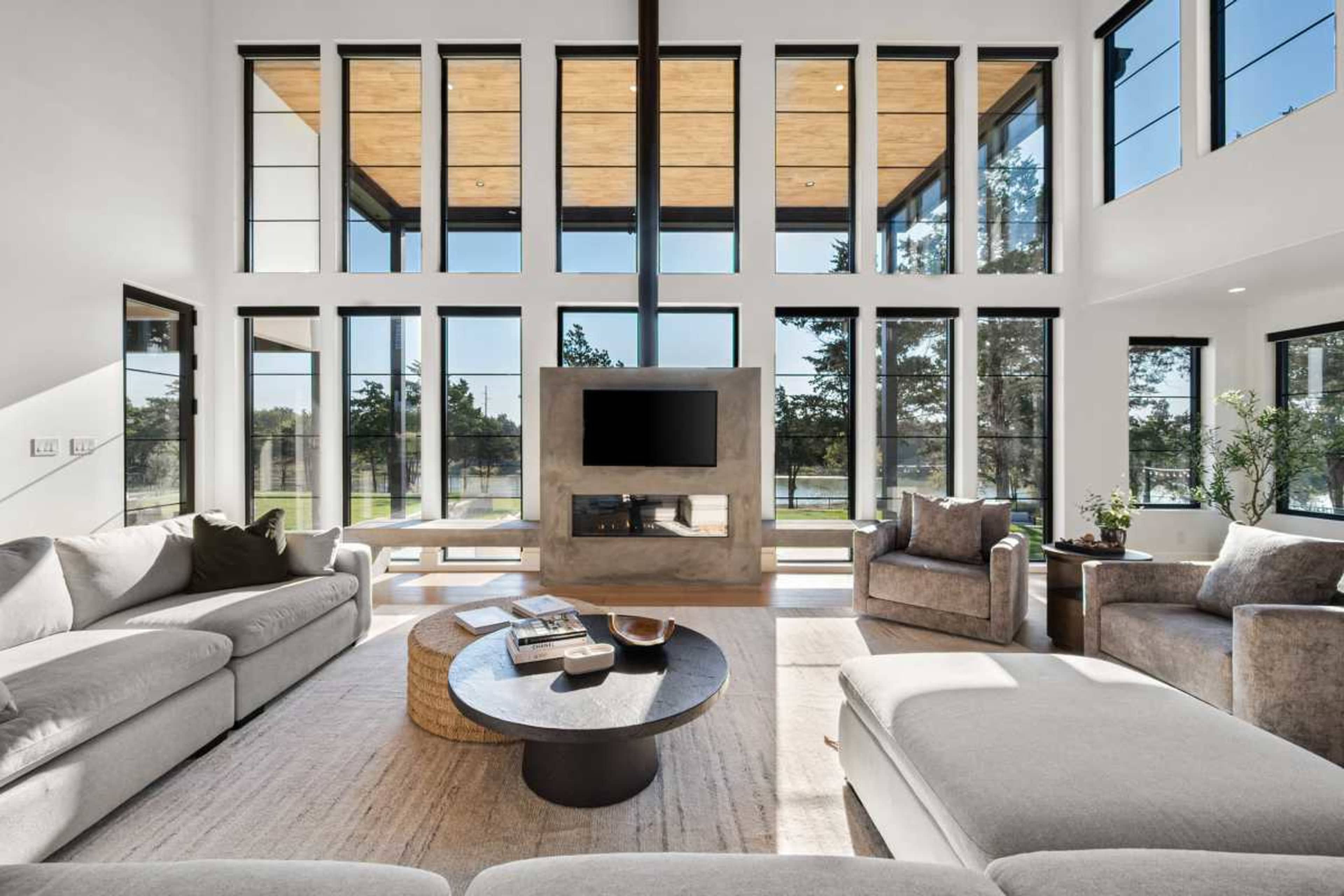 A bright and spacious living room features large windows, a central circular coffee table, and comfortable seating arranged around a modern fireplace.