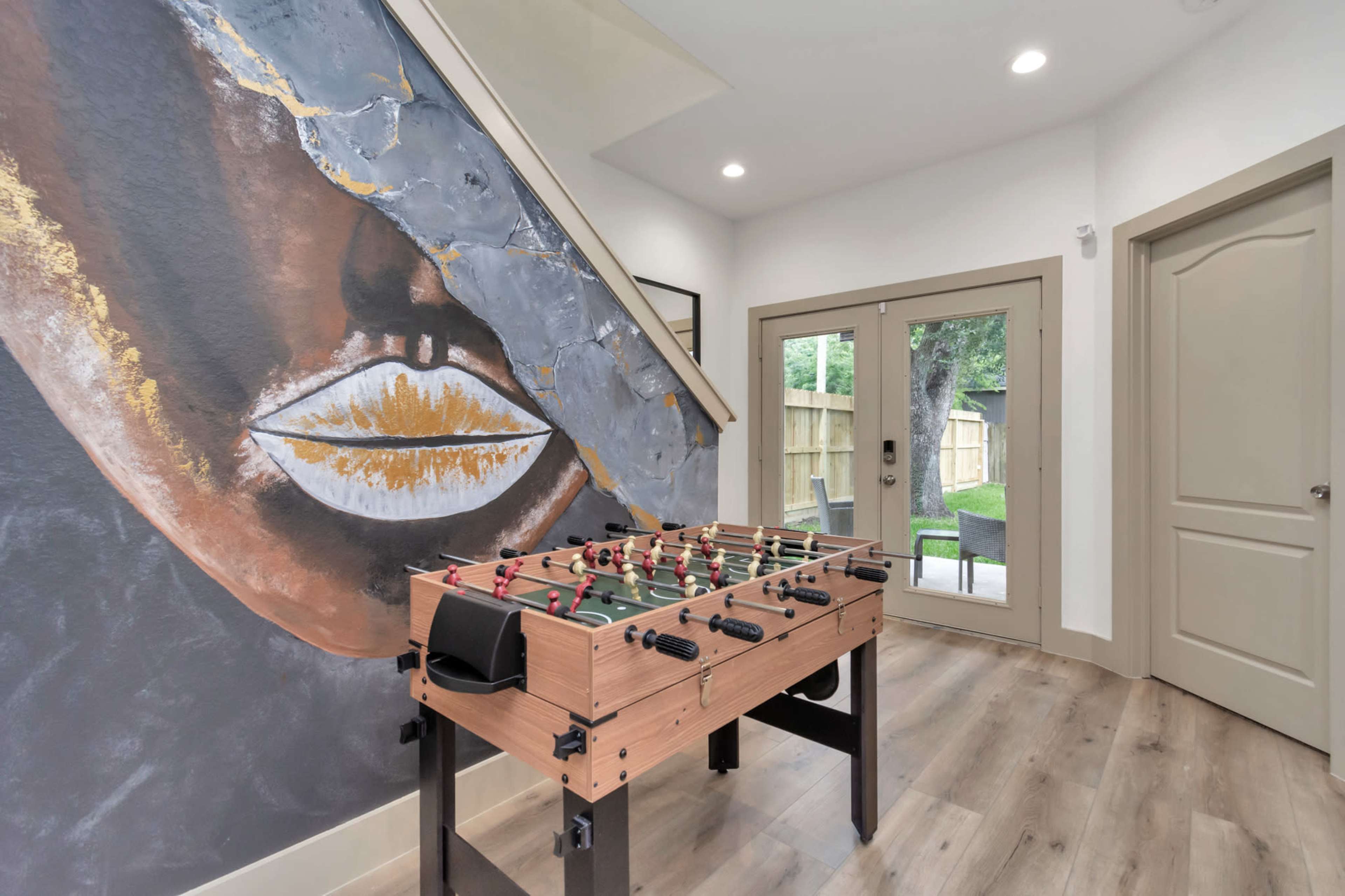 A foosball table is positioned near a staircase against a wall featuring a large mural of a face with striking gold accents.