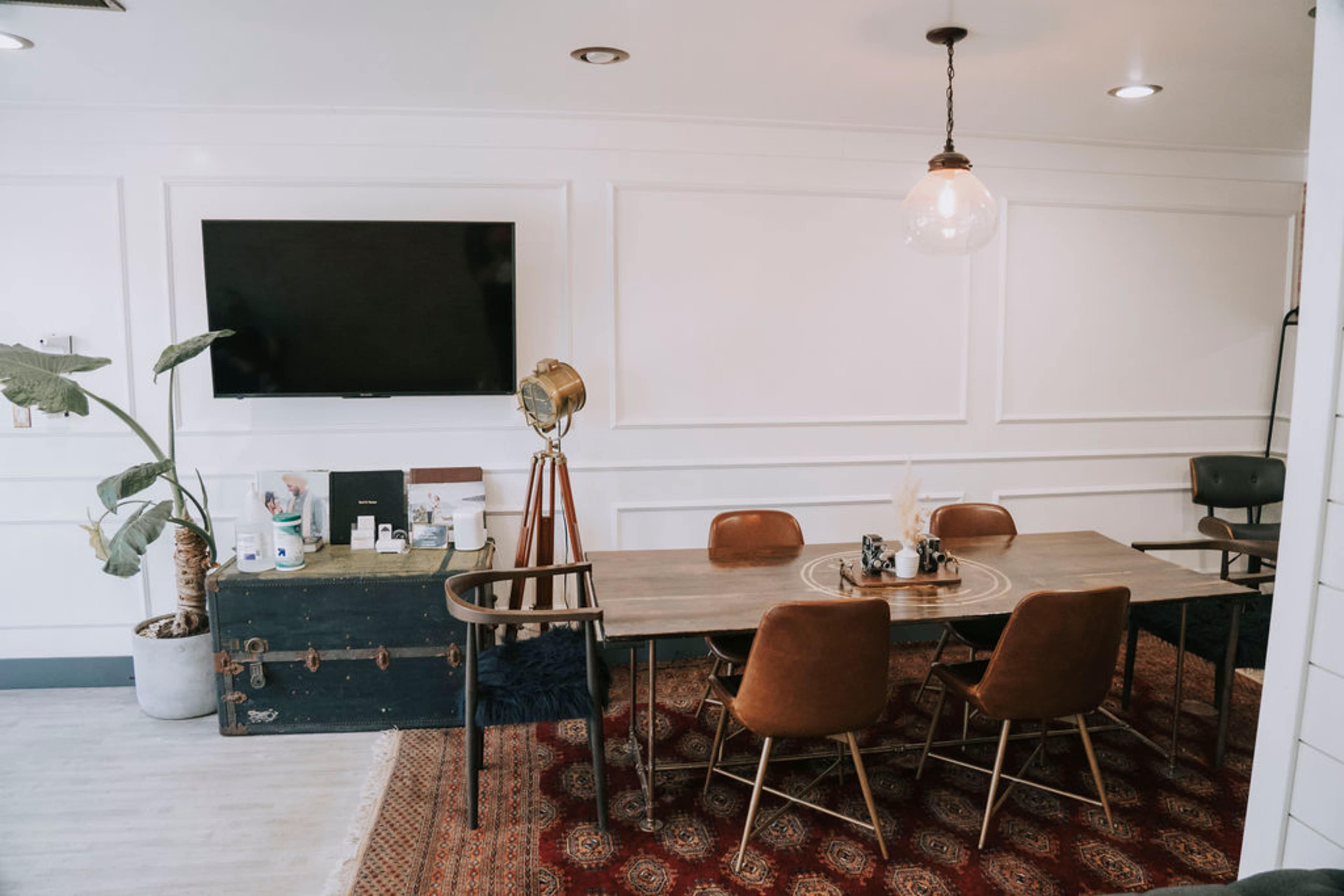 A modern dining area features a large table with brown leather chairs, a decorative rug, a television on the wall, and a standing lamp in the corner.