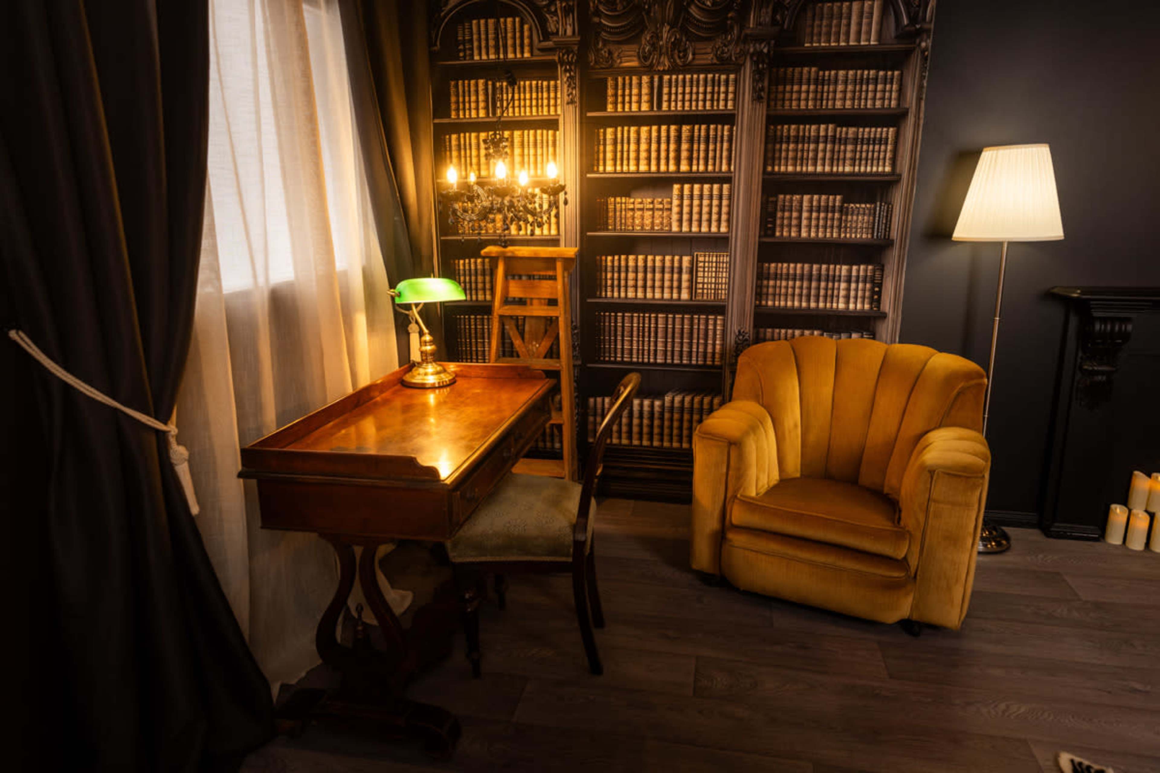 The image shows a cozy reading nook featuring a wooden desk with a chair, a yellow armchair, a lamp, and a bookshelf filled with books.
