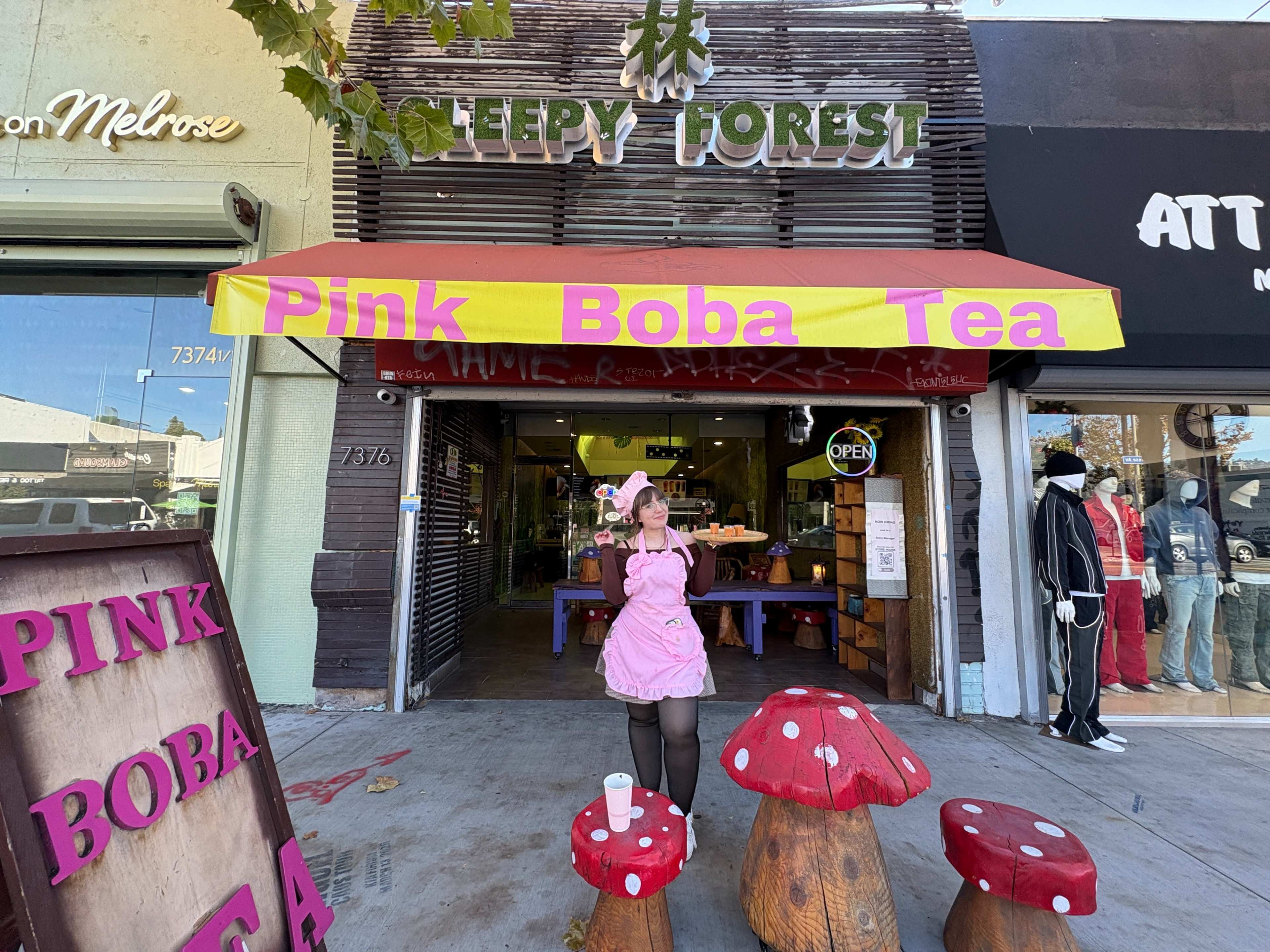 A woman in a pink costume poses in front of a shop called "Sleepy Forest," which has a bright yellow awning that reads "Pink Boba Tea."