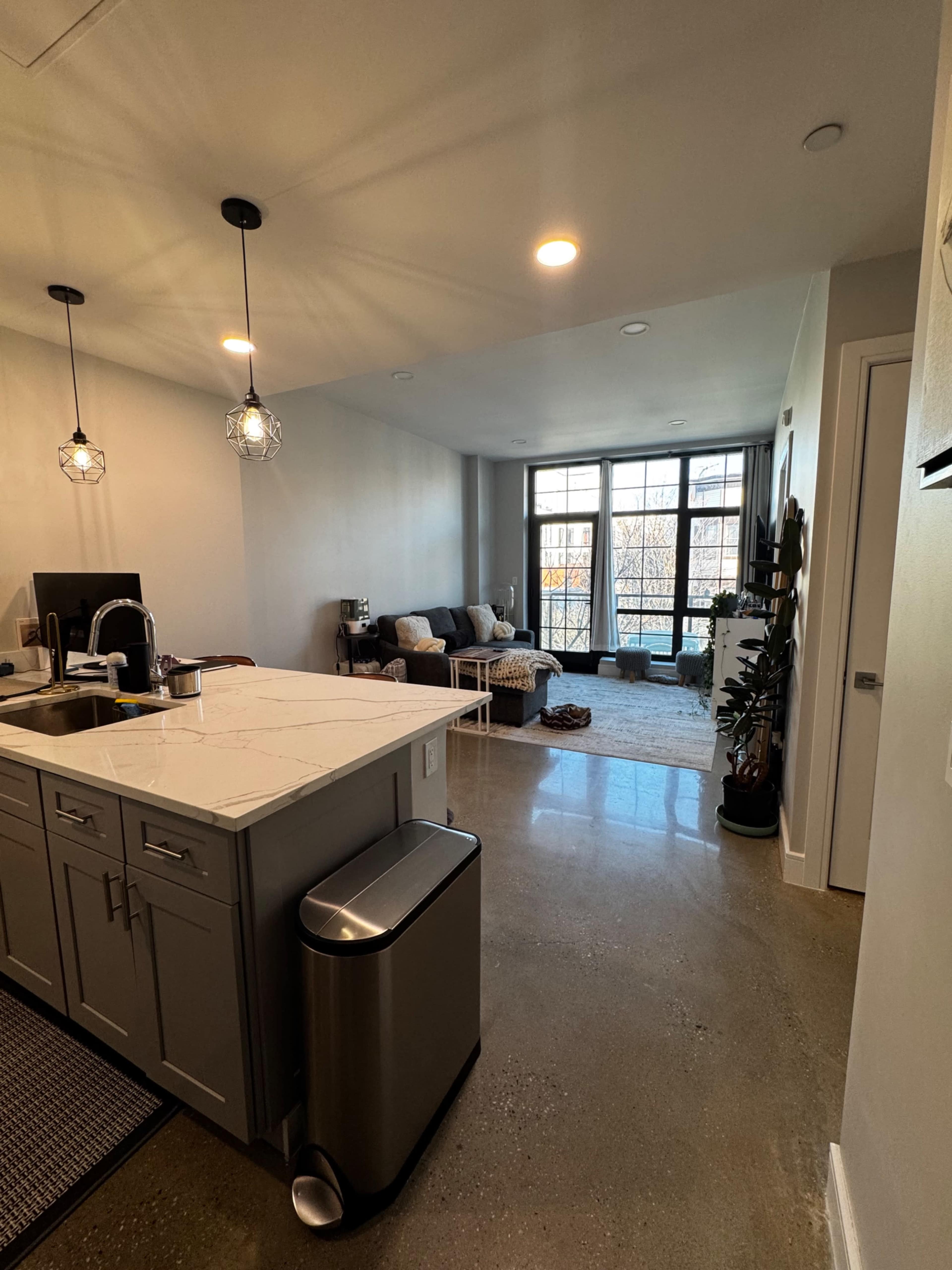 The image shows a modern kitchen with a marble countertop leading into a living area that features a couch, rug, and large windows.