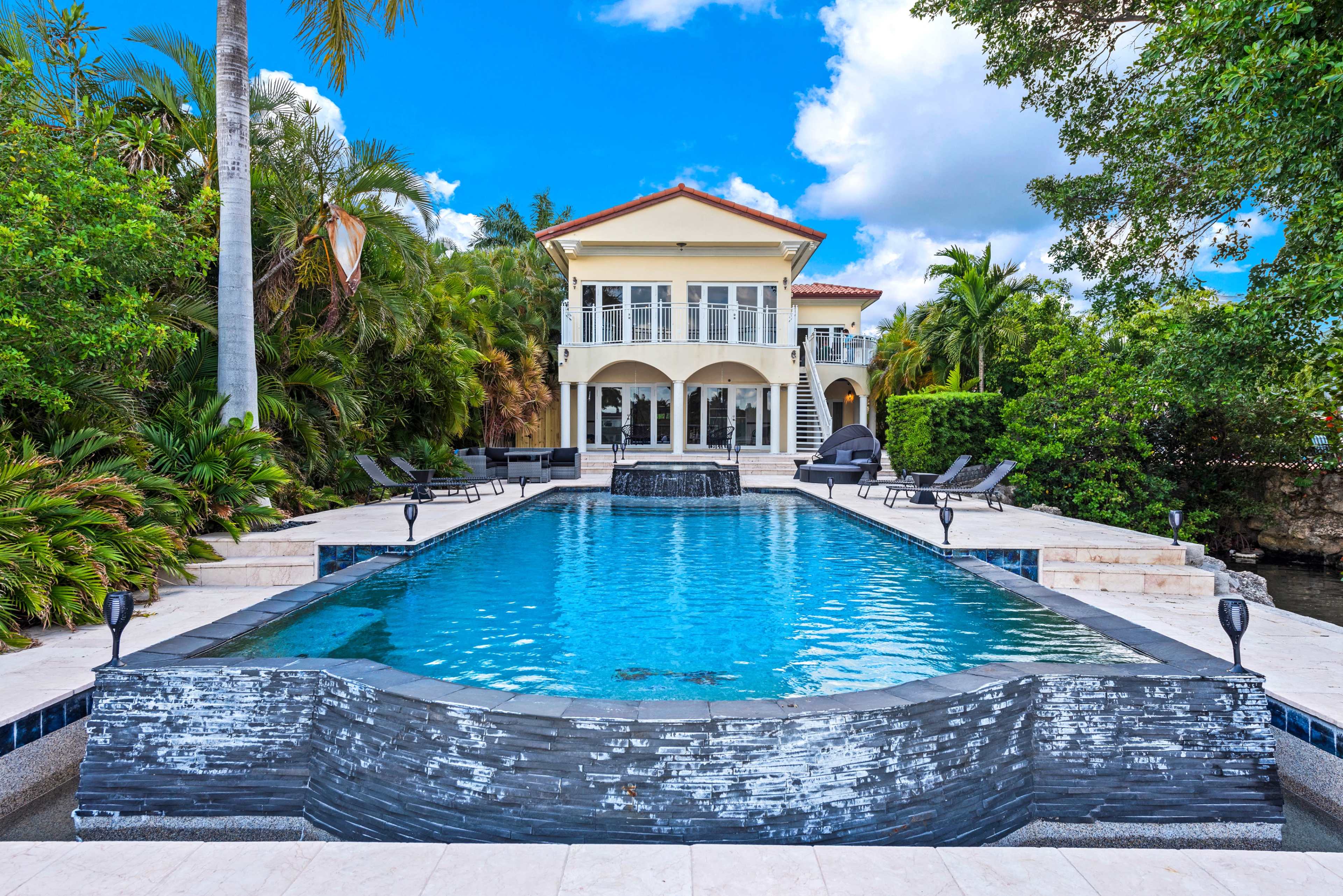 A spacious swimming pool with a waterfall feature in front of a multi-story house surrounded by tropical landscaping.