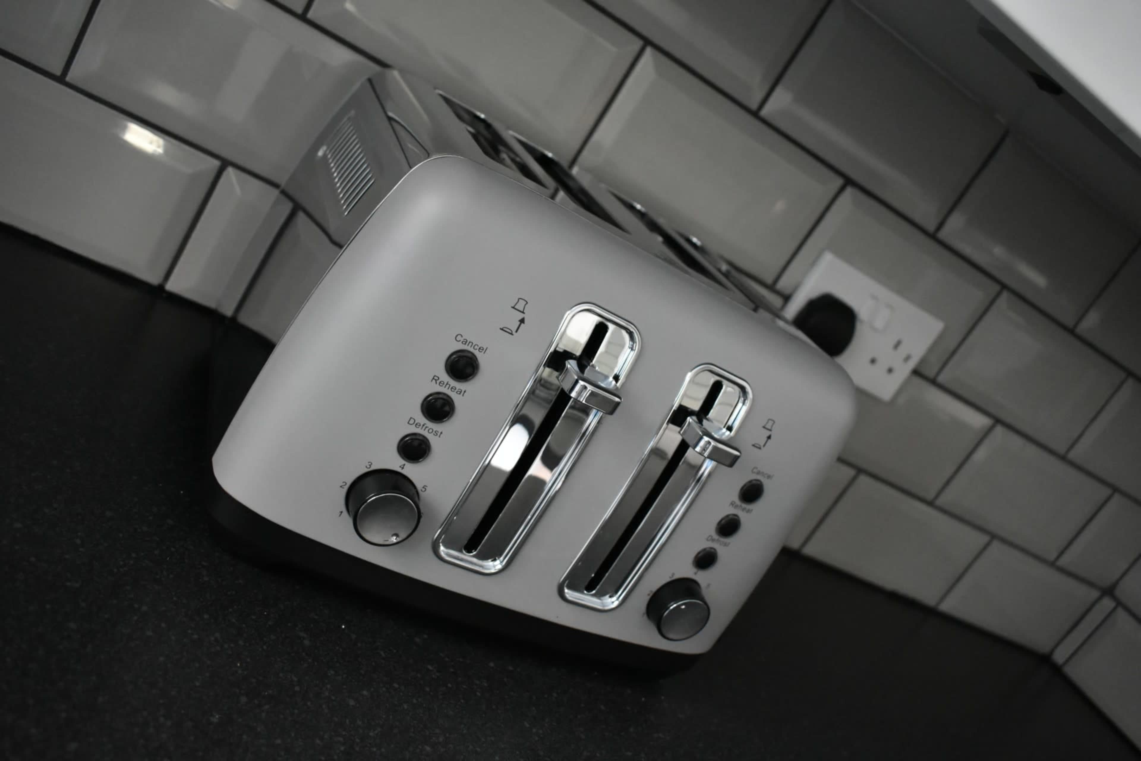 A silver four-slot toaster with adjustable settings on a black countertop against a tiled wall.