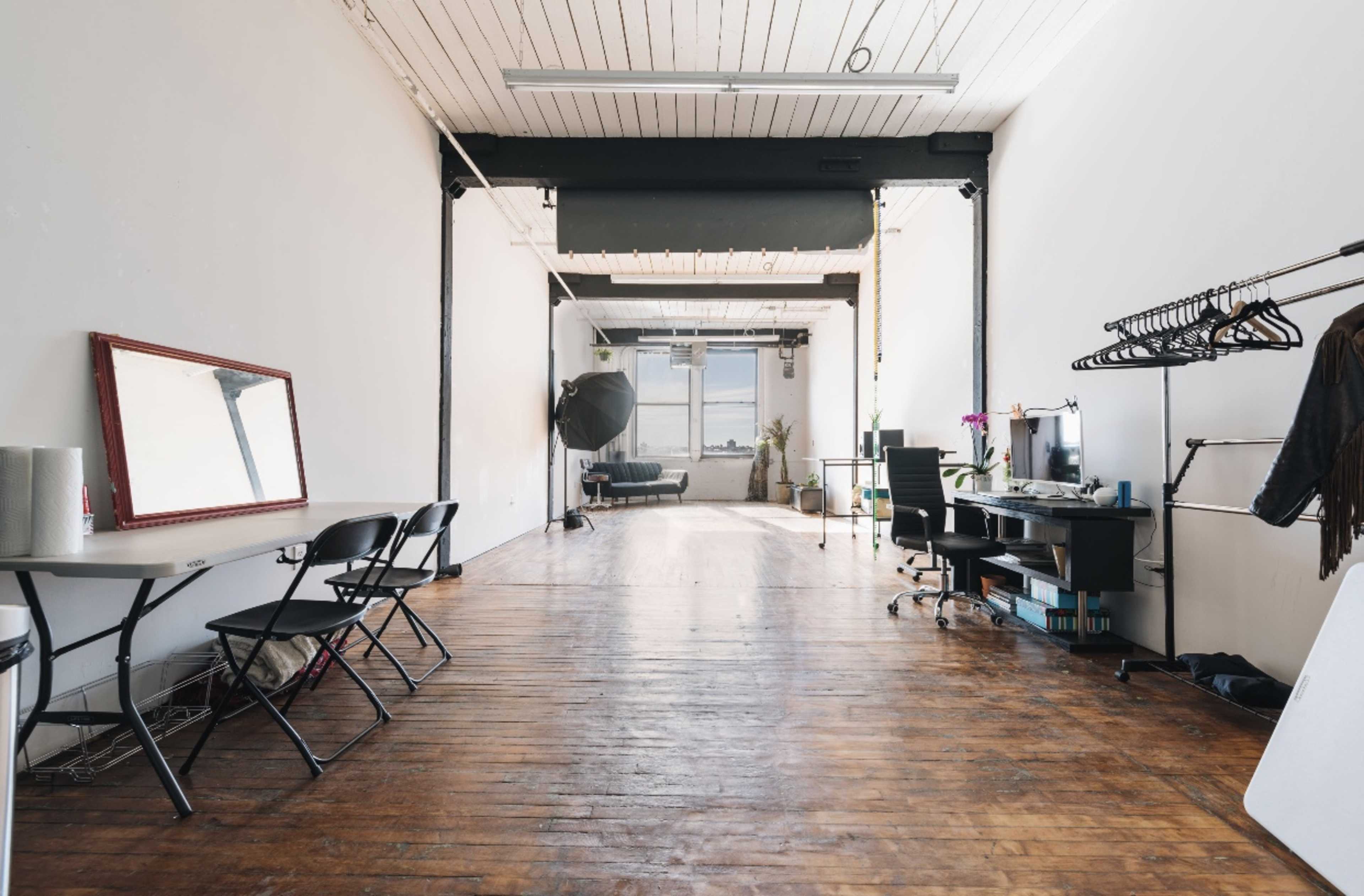 A spacious, minimalist studio with wooden flooring, a large window, a seating area, and several functional furnishings, including tables and clothing racks.