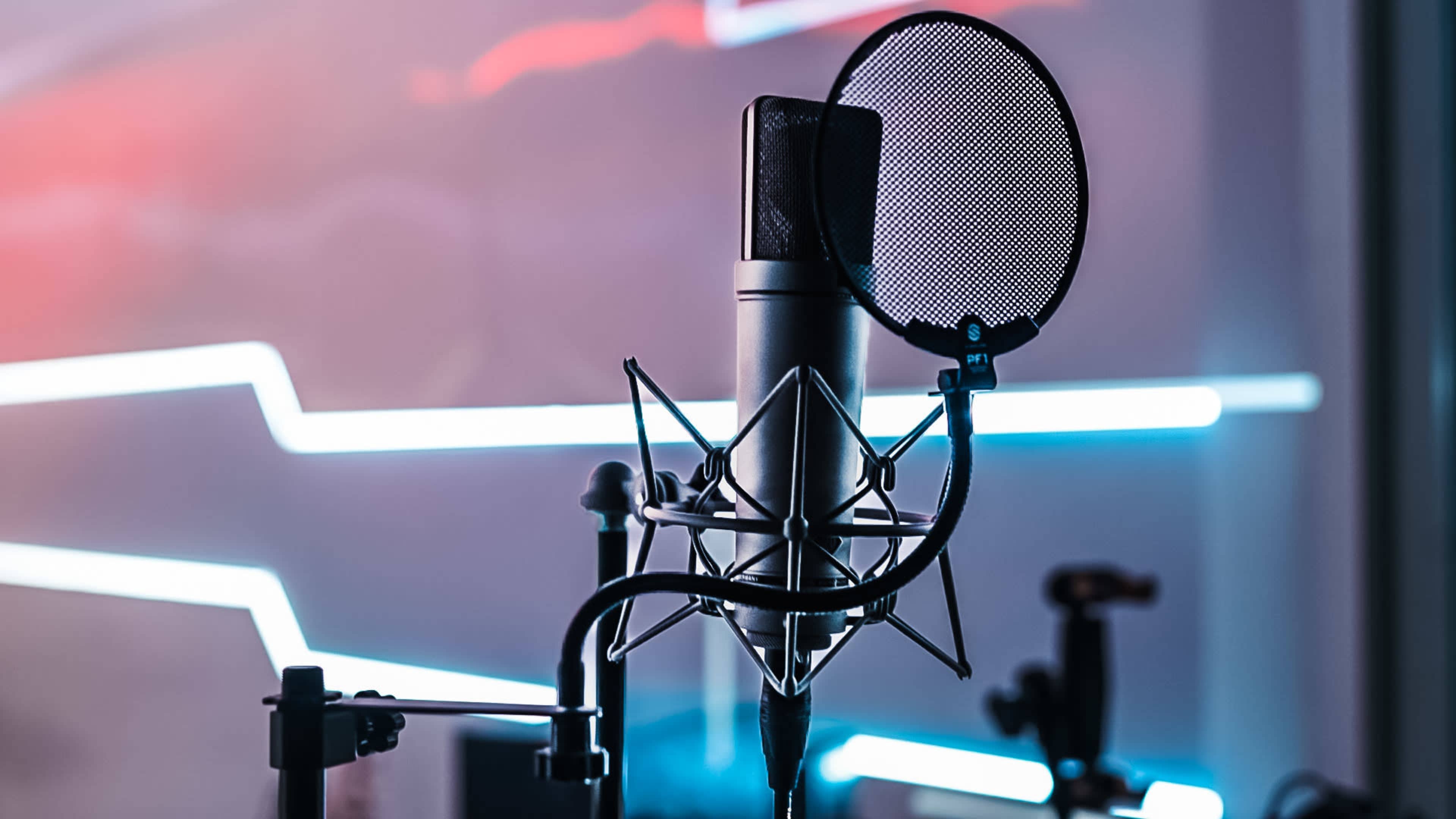 A studio microphone mounted on a stand with a pop filter in front of it, set against a backdrop of colored lights.