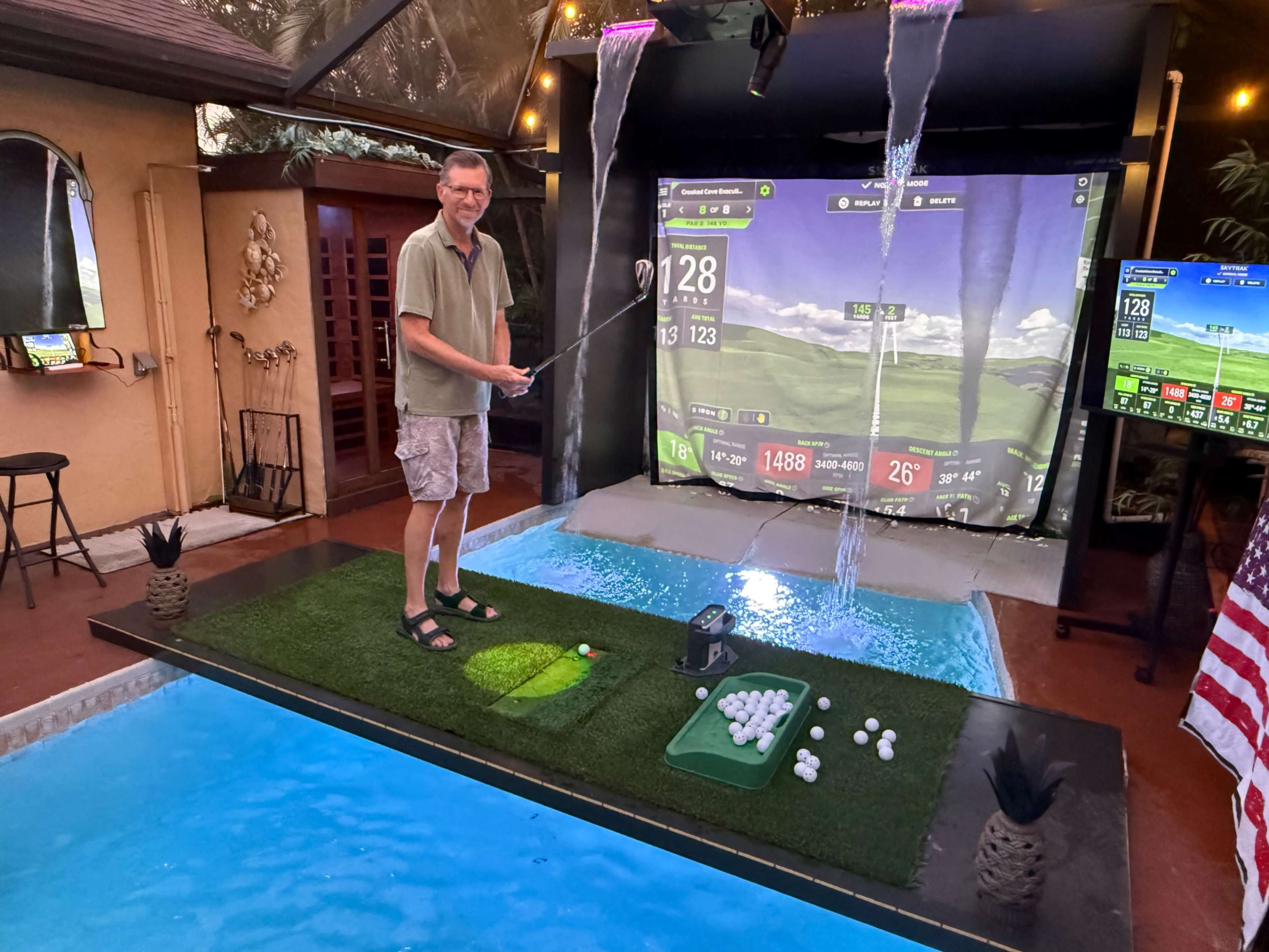 A man stands on an artificial green in a home golf simulator room, preparing to hit a golf ball while a large screen displays a golf course and water features are visible in the background.