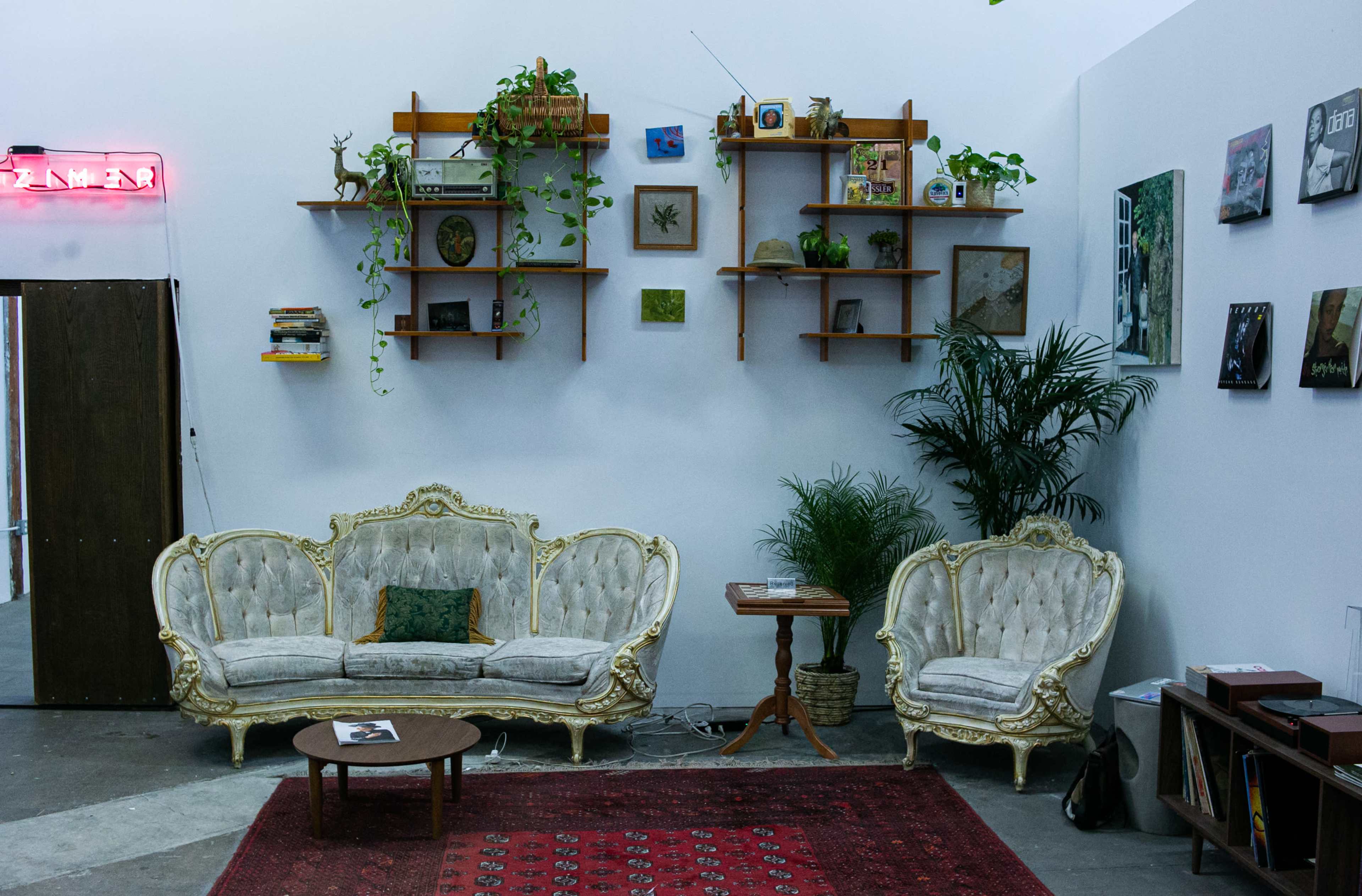 The image shows a vintage-style seating area with two ornate sofas, a small table, and shelves adorned with plants and decor items against a white wall.