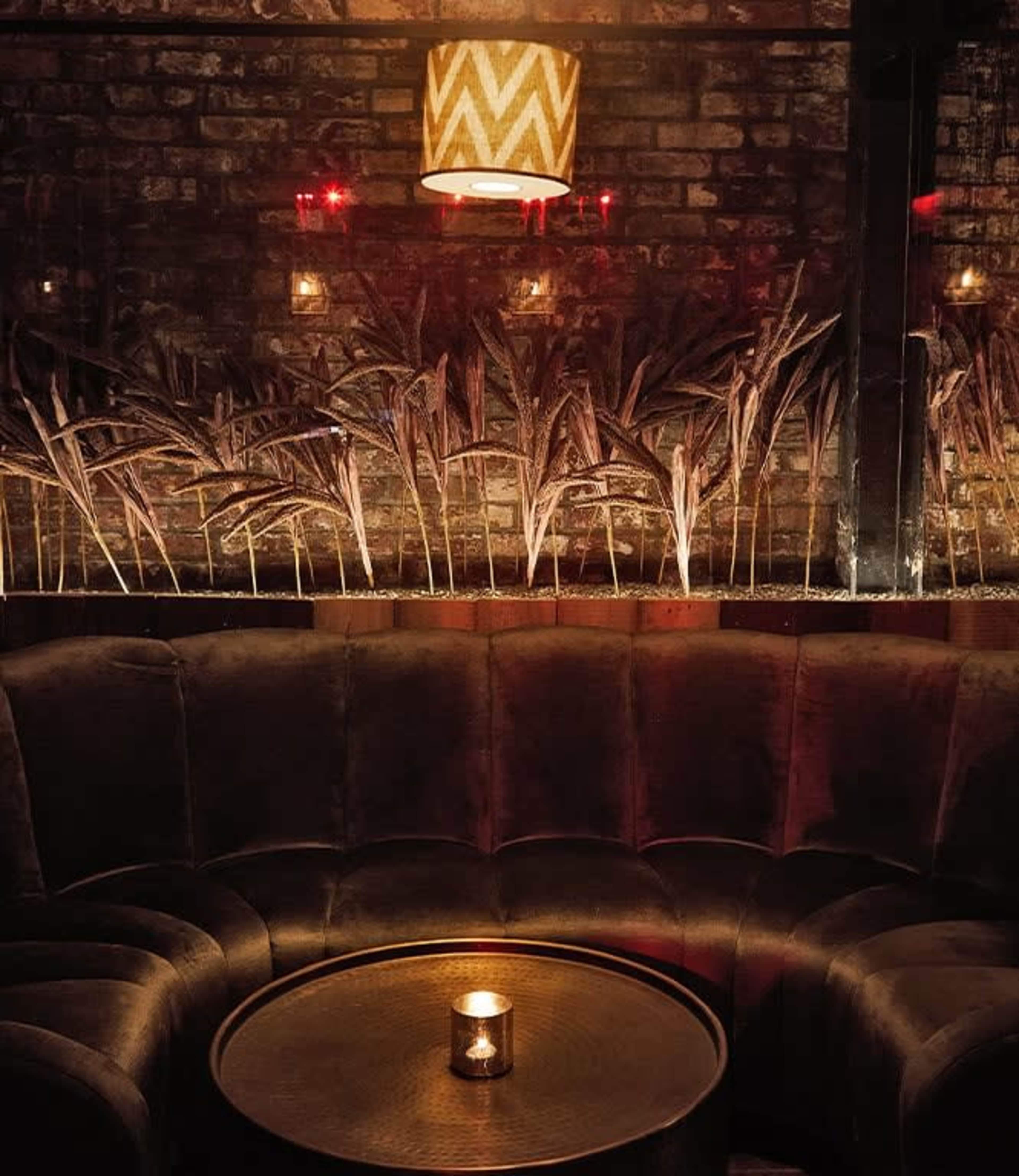 A cozy seating area features a curved velvet couch, a round table with a candle, and a backdrop of illuminated plants against a brick wall.