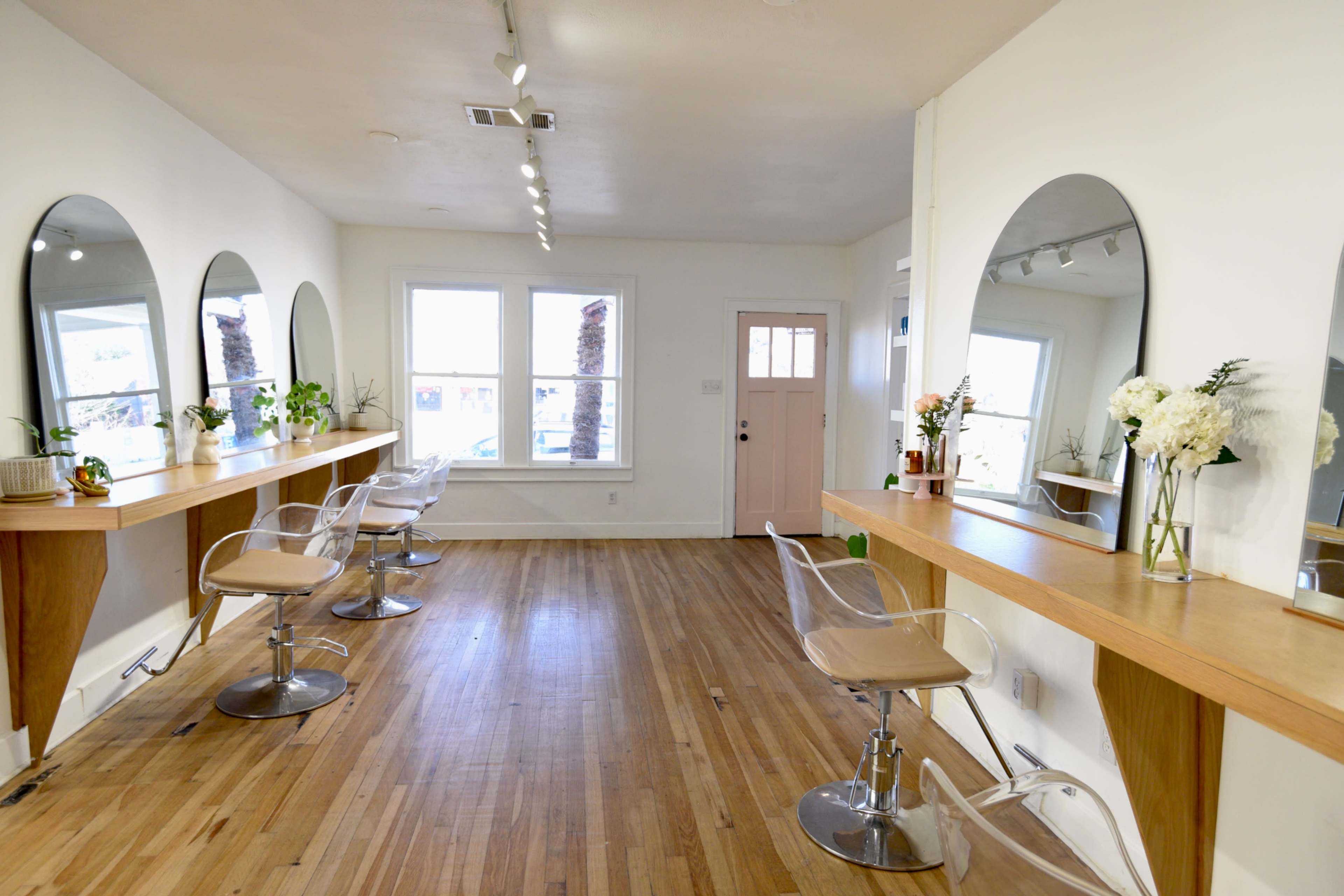 A well-lit salon with three mirrored styling stations and clear chairs, featuring wooden counters and large windows.