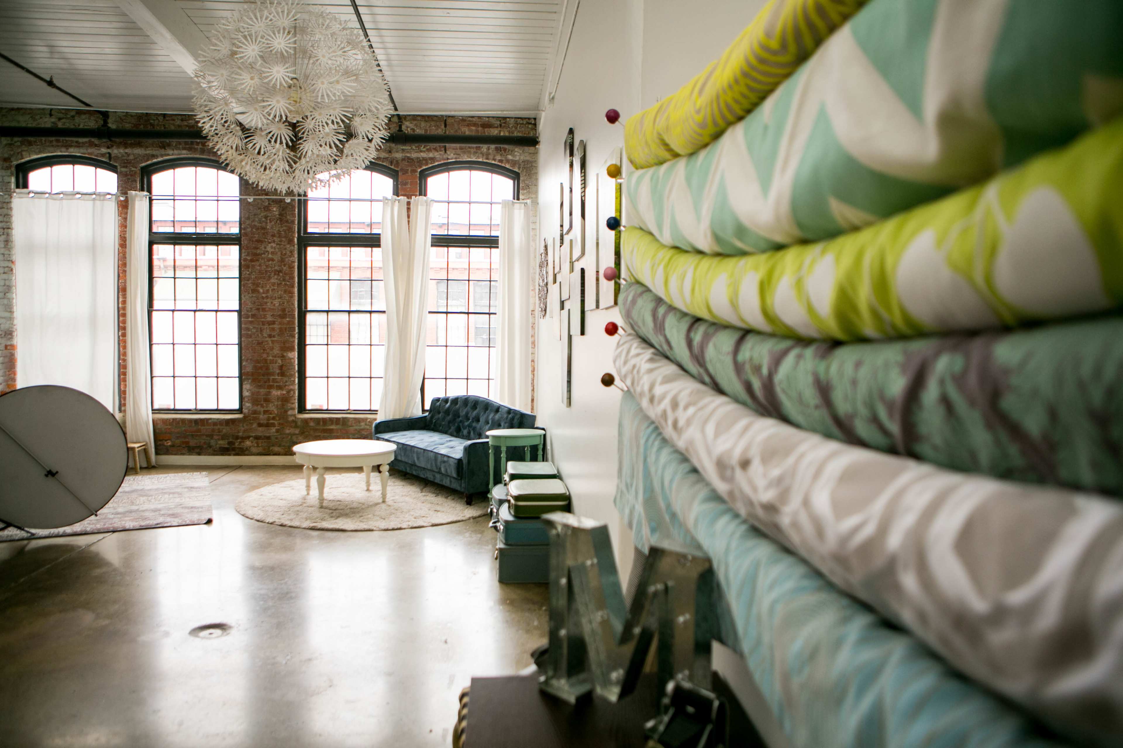 A bright, spacious interior with large windows, a blue couch, and a stack of colorful fabric rolls against the wall.