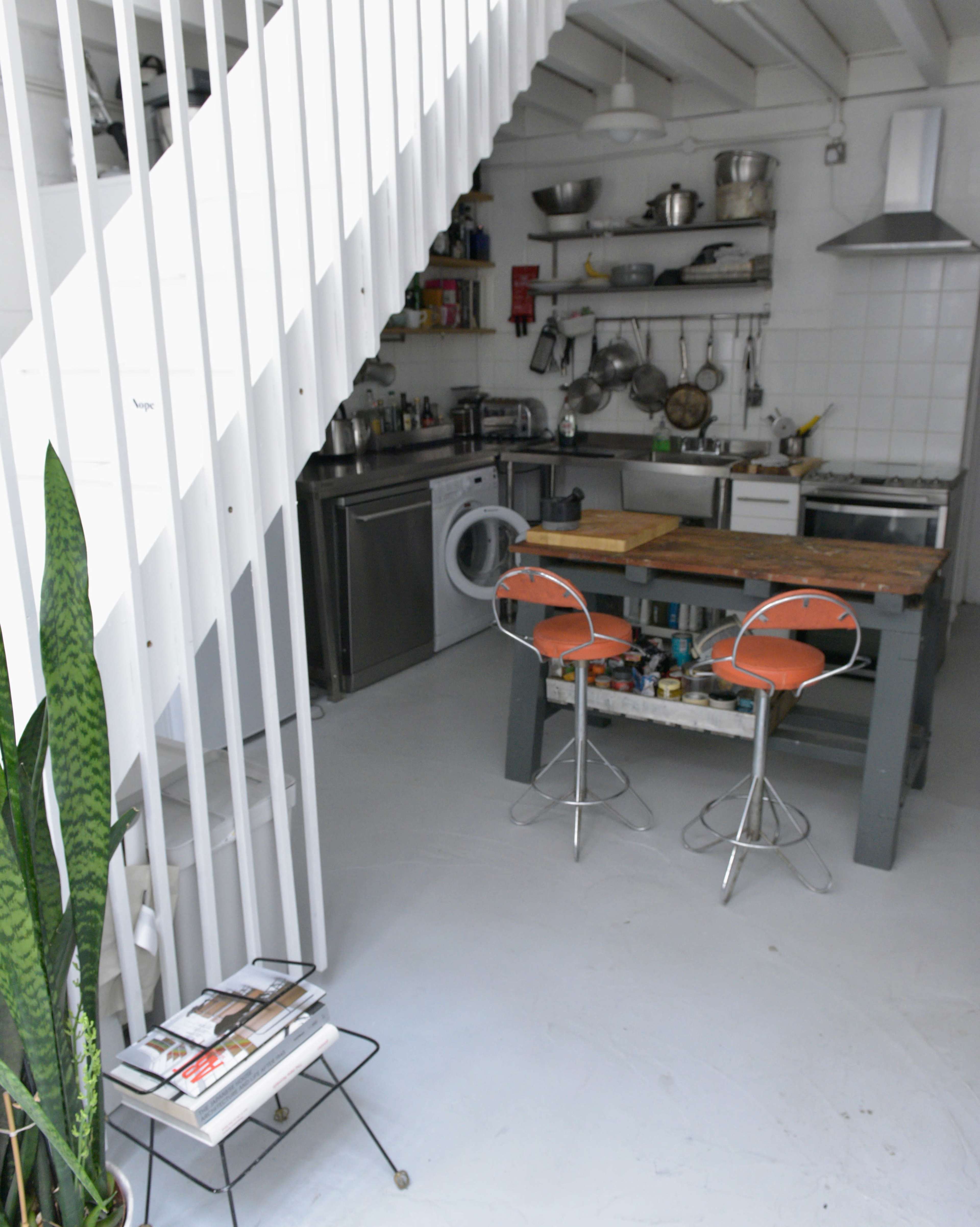 A modern kitchen with a staircase, featuring stainless steel appliances, a wooden table with two orange bar stools, and shelves filled with kitchenware.